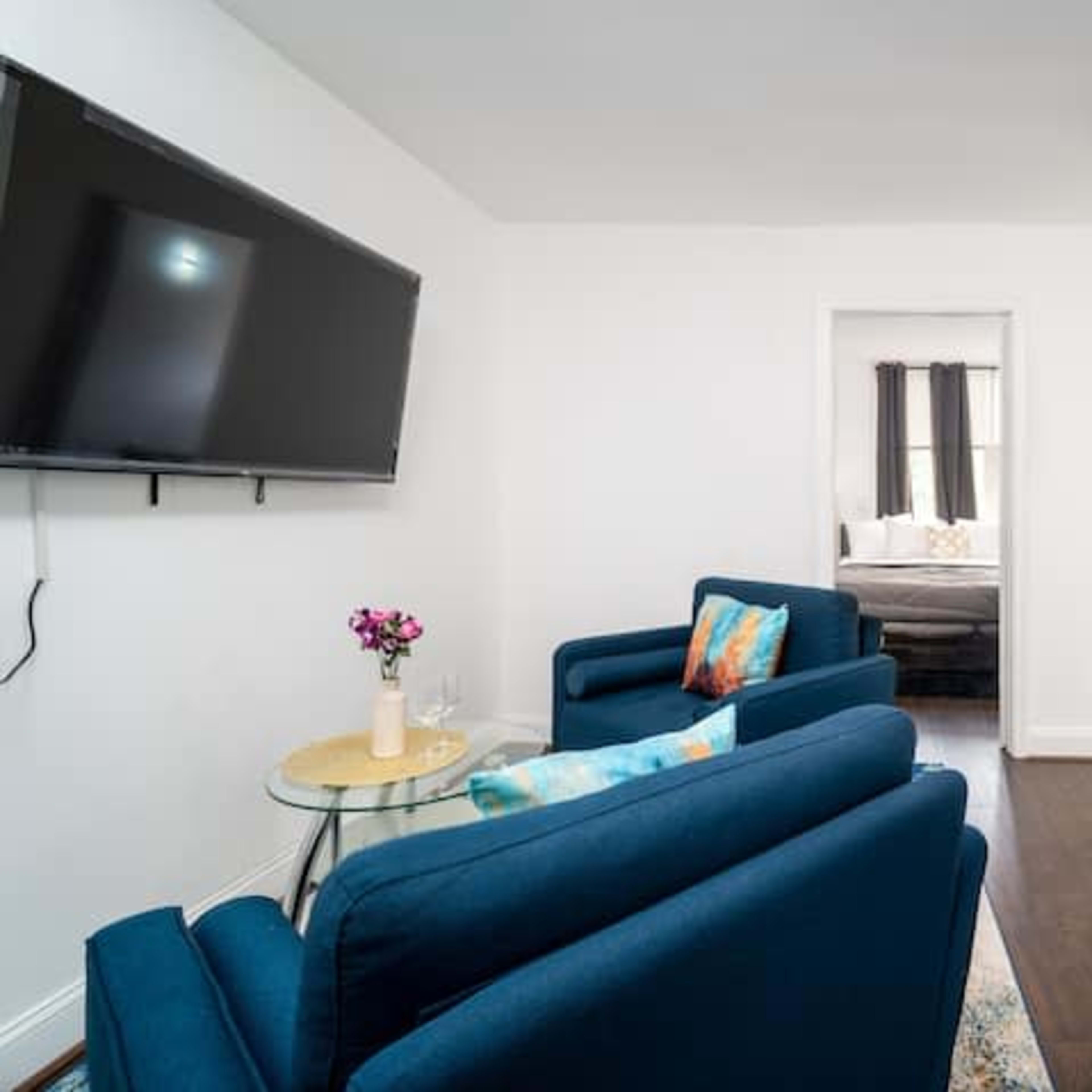 A modern living space featuring two blue chairs, a small glass table with flowers, a wall-mounted TV, and a doorway leading to a bedroom and a laundry area with a washing machine.
