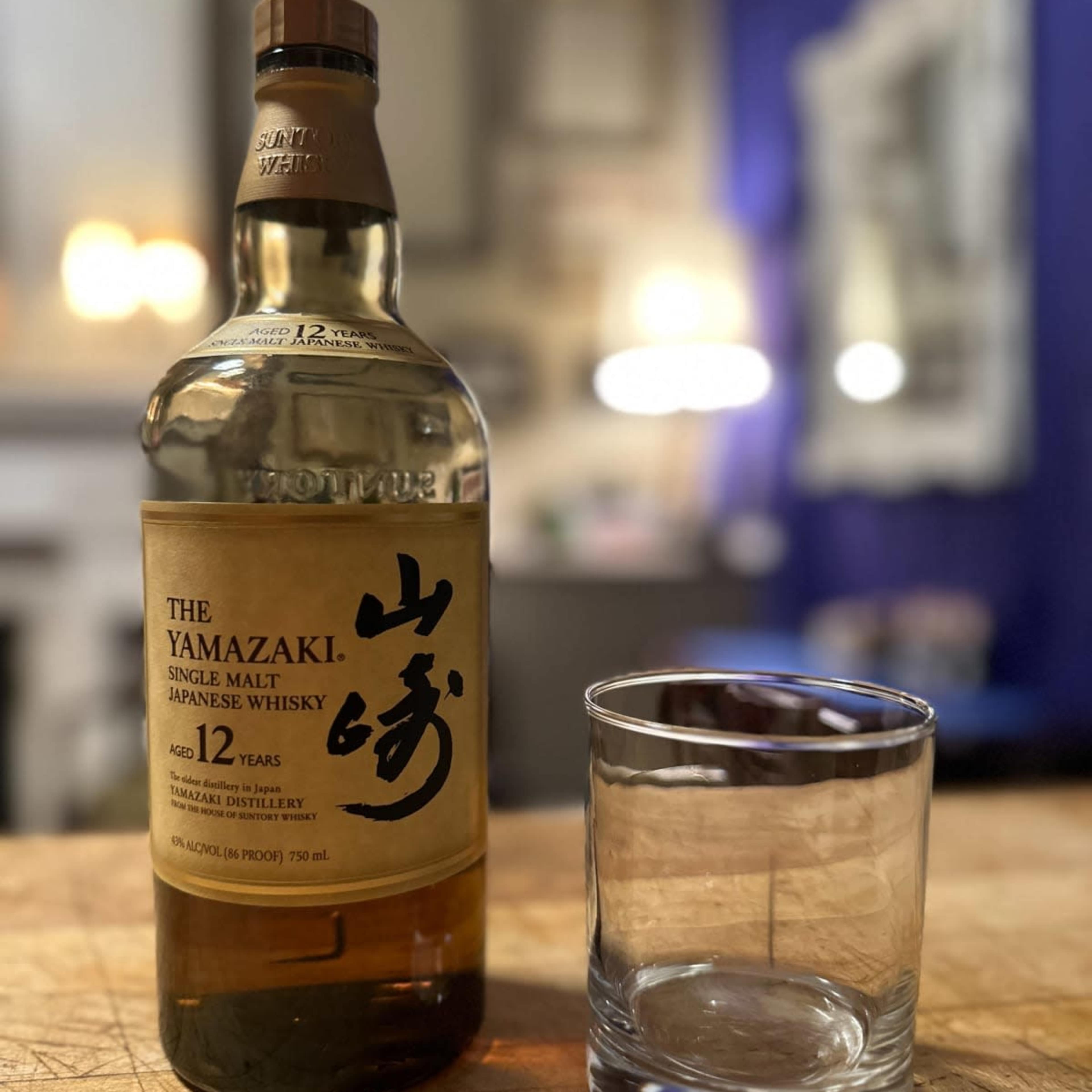A bottle of Yamazaki 12 Year Old Single Malt Japanese Whisky stands next to an empty glass on a wooden countertop.