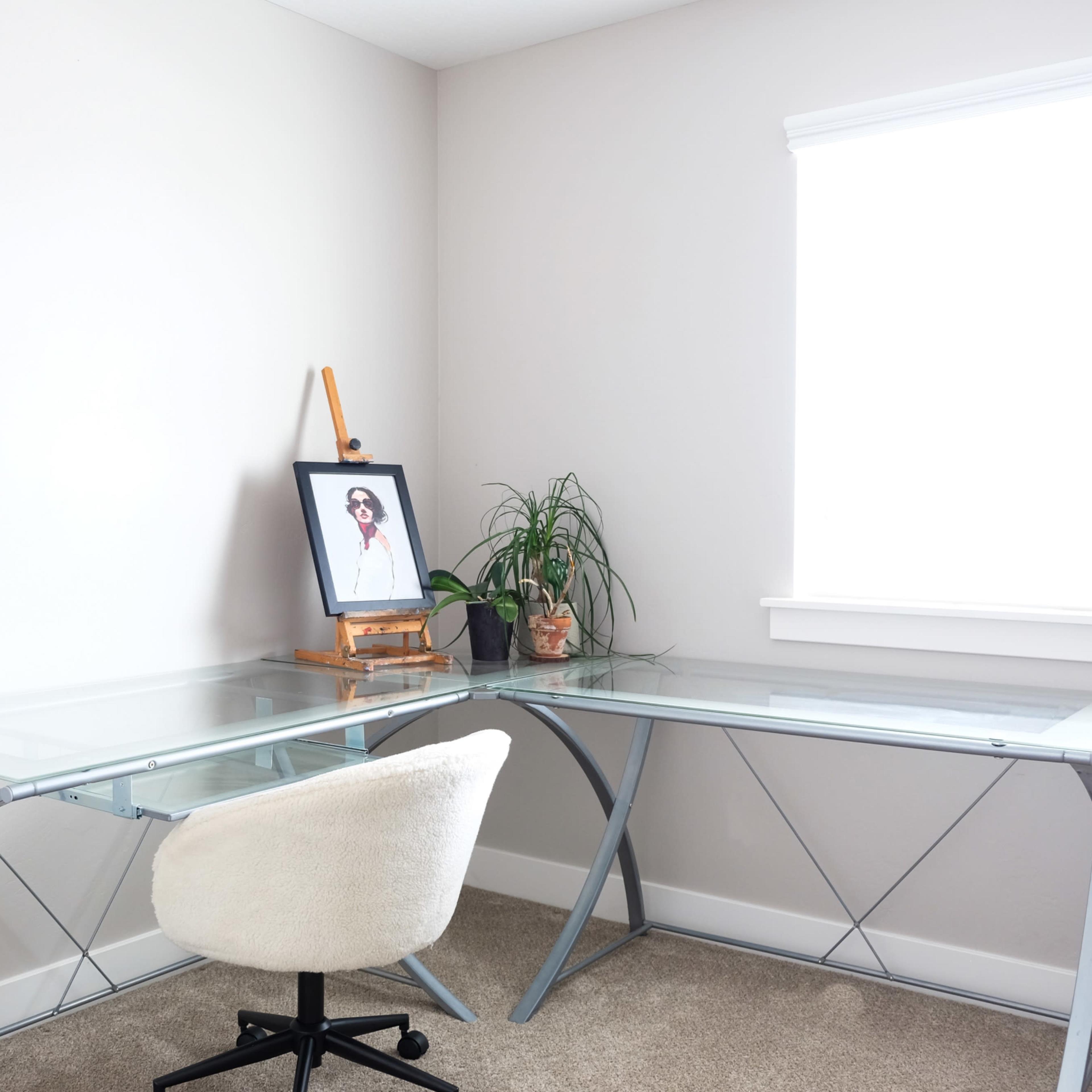 A glass-top desk with a chair is positioned in a corner, featuring a plant and a framed picture on an easel.