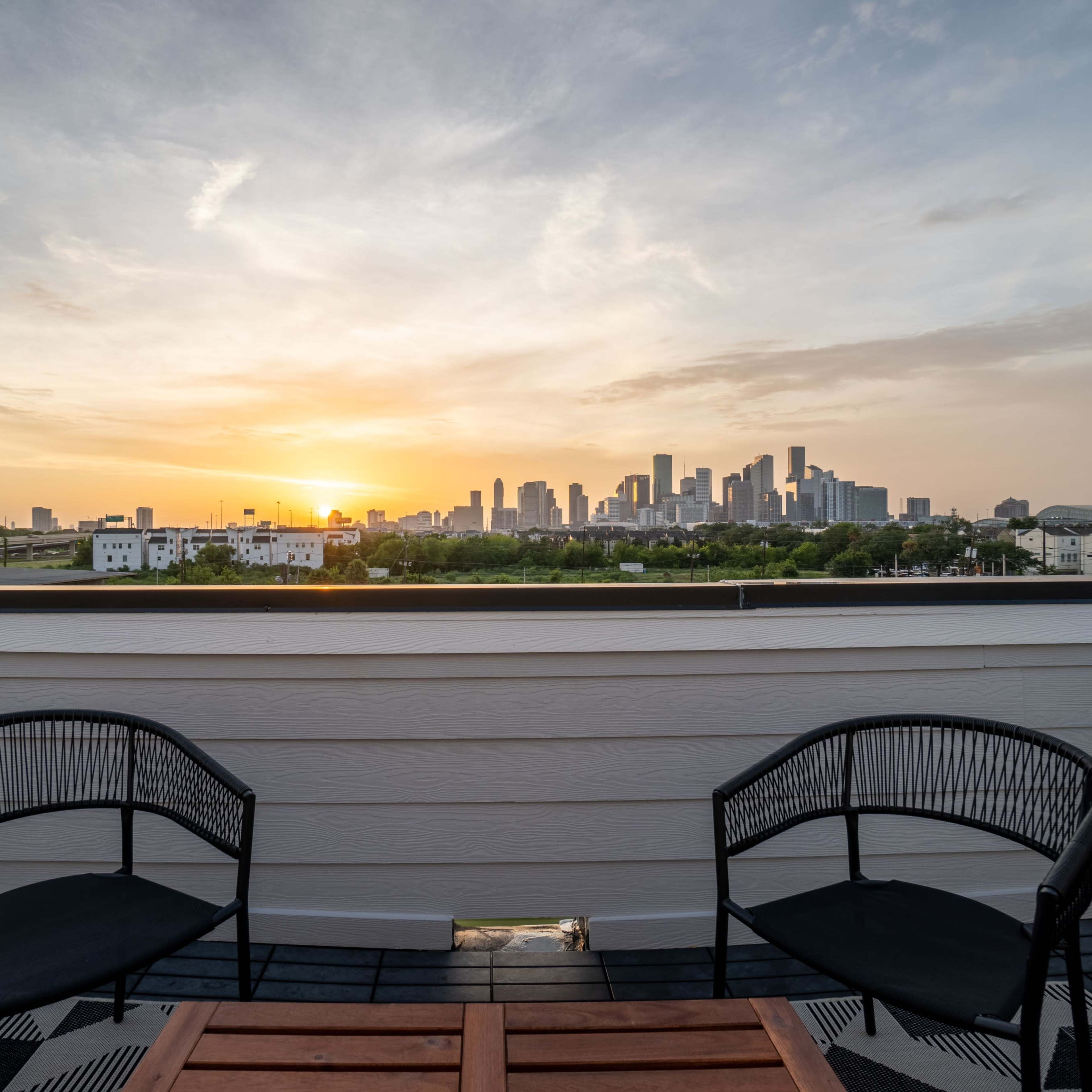 A rooftop view of a city skyline at sunset, framed by two chairs and a table on the balcony.