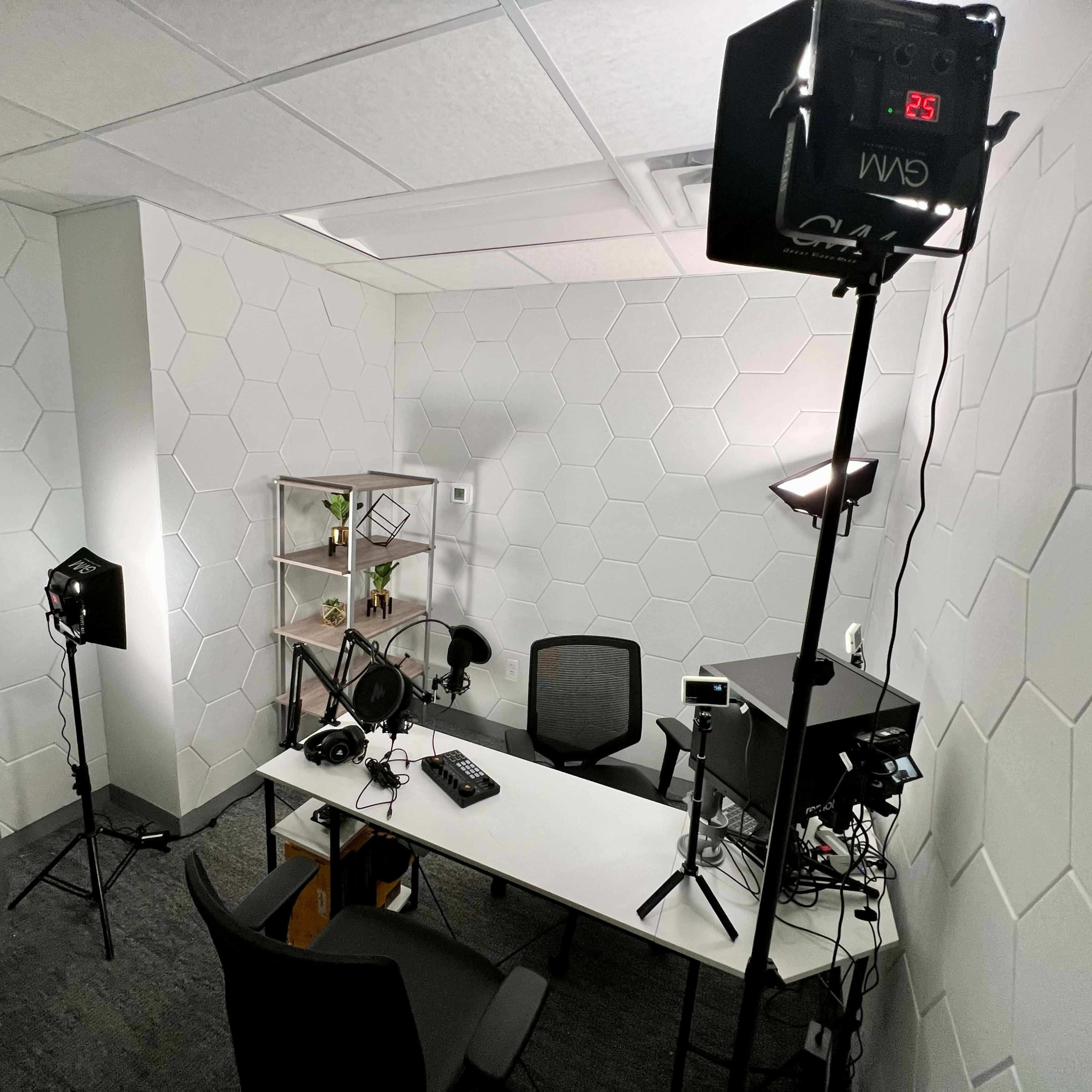 A small studio setup with a hexagon-patterned wall, a white desk with audio equipment, two chairs, and lighting fixtures placed around the space.