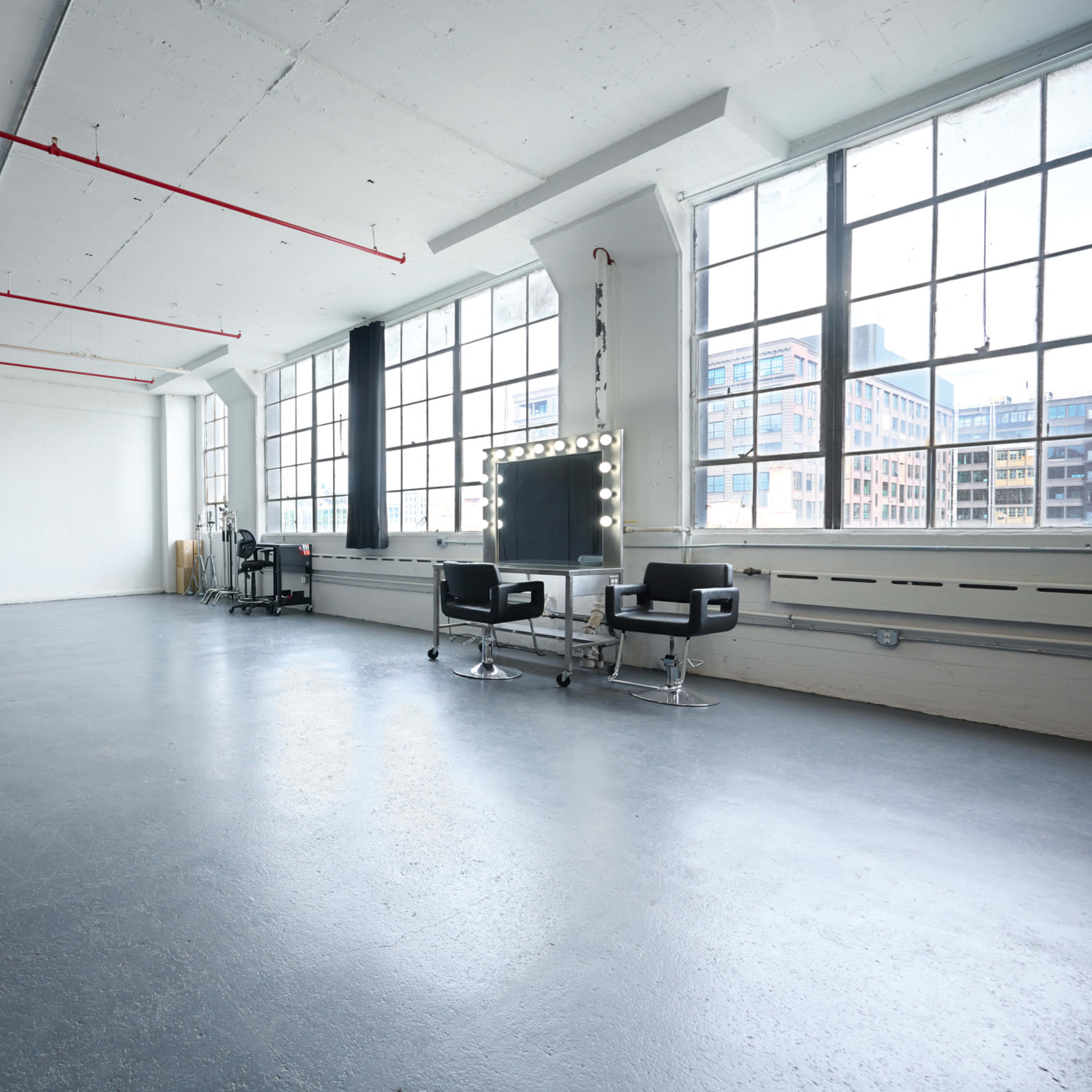 A spacious studio with large windows, black curtains on one side, and two styling chairs next to a mirror with lights.