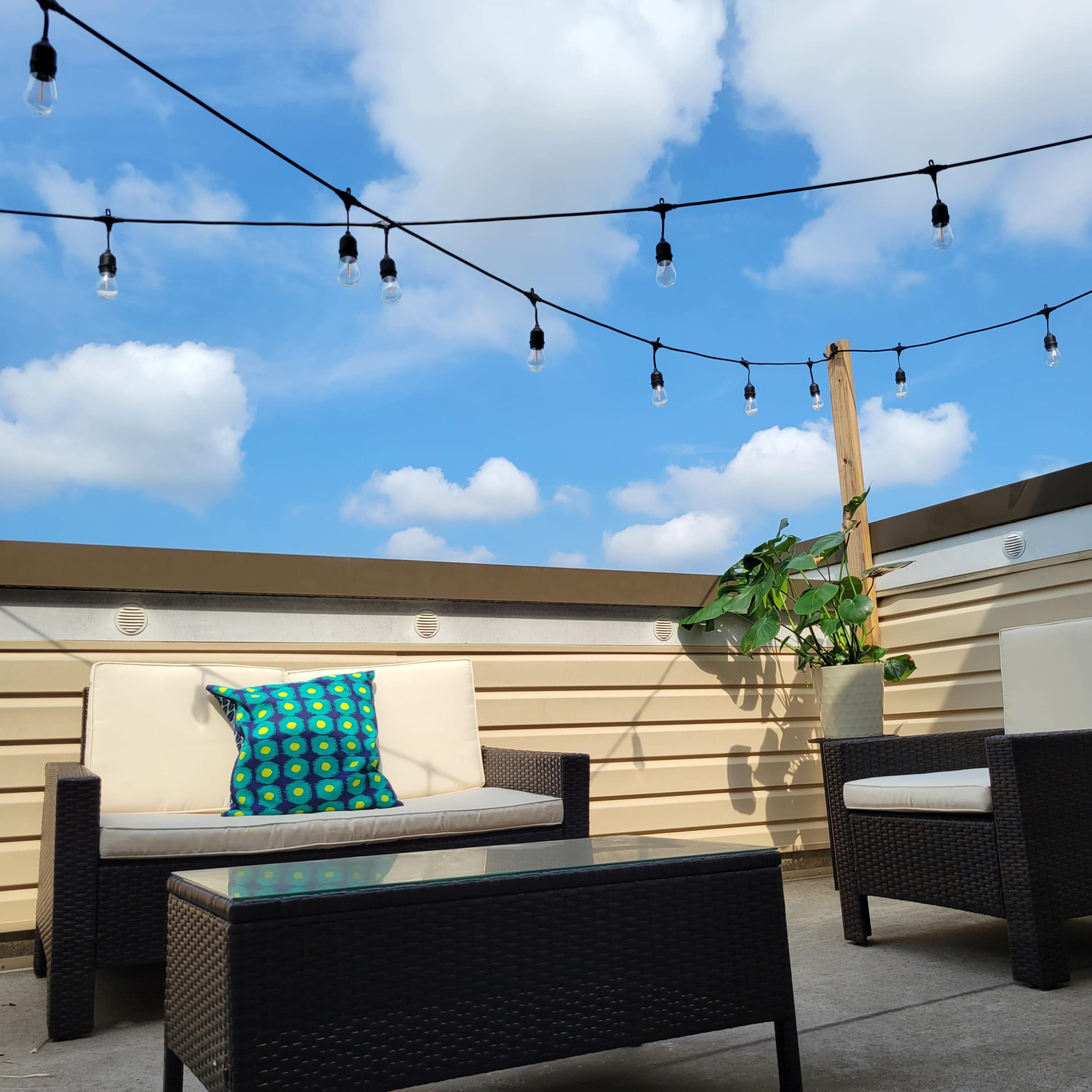 A rooftop patio features two chairs and a table, with a colorful pillow on one chair and string lights overhead against a backdrop of blue sky and clouds.