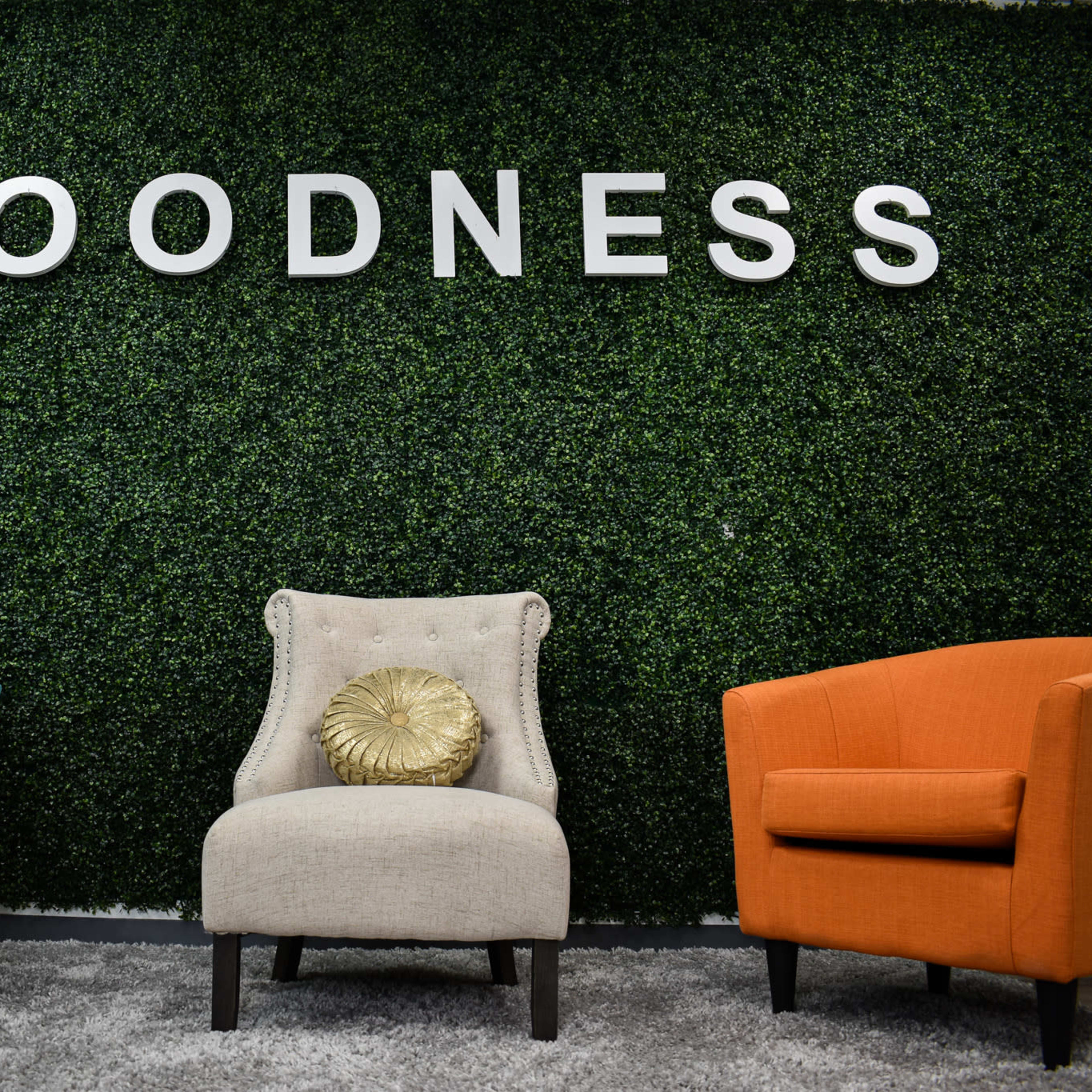 Two armchairs, one turquoise and one orange, facing a beige chair with a decorative cushion, against a green wall with the word "GOODNESS" prominently displayed.