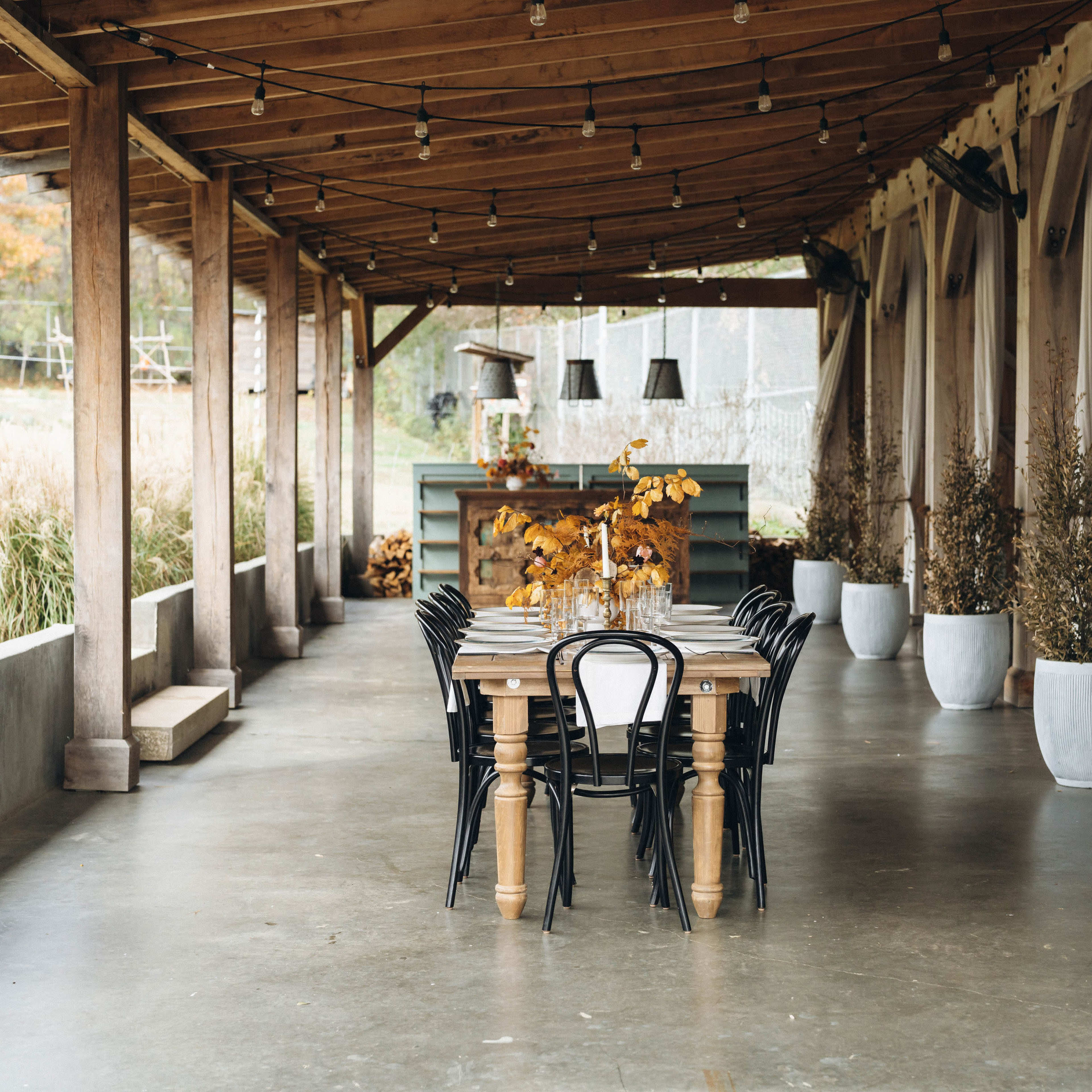 A long wooden table is set with black chairs beneath a covered porch adorned with hanging lights and potted plants.