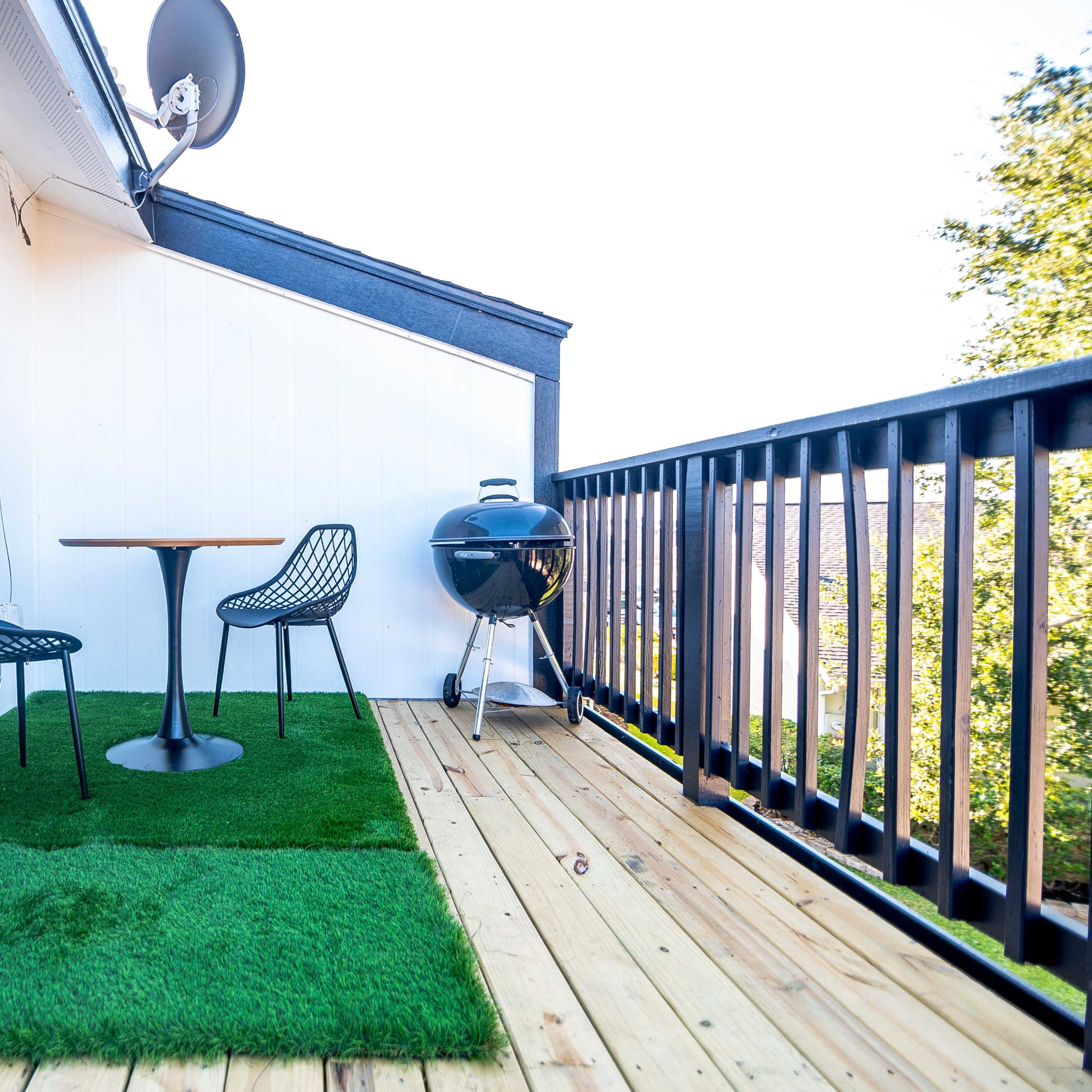 A small balcony features a round table and two chairs on artificial grass, alongside a barbecue grill and a portable speaker.