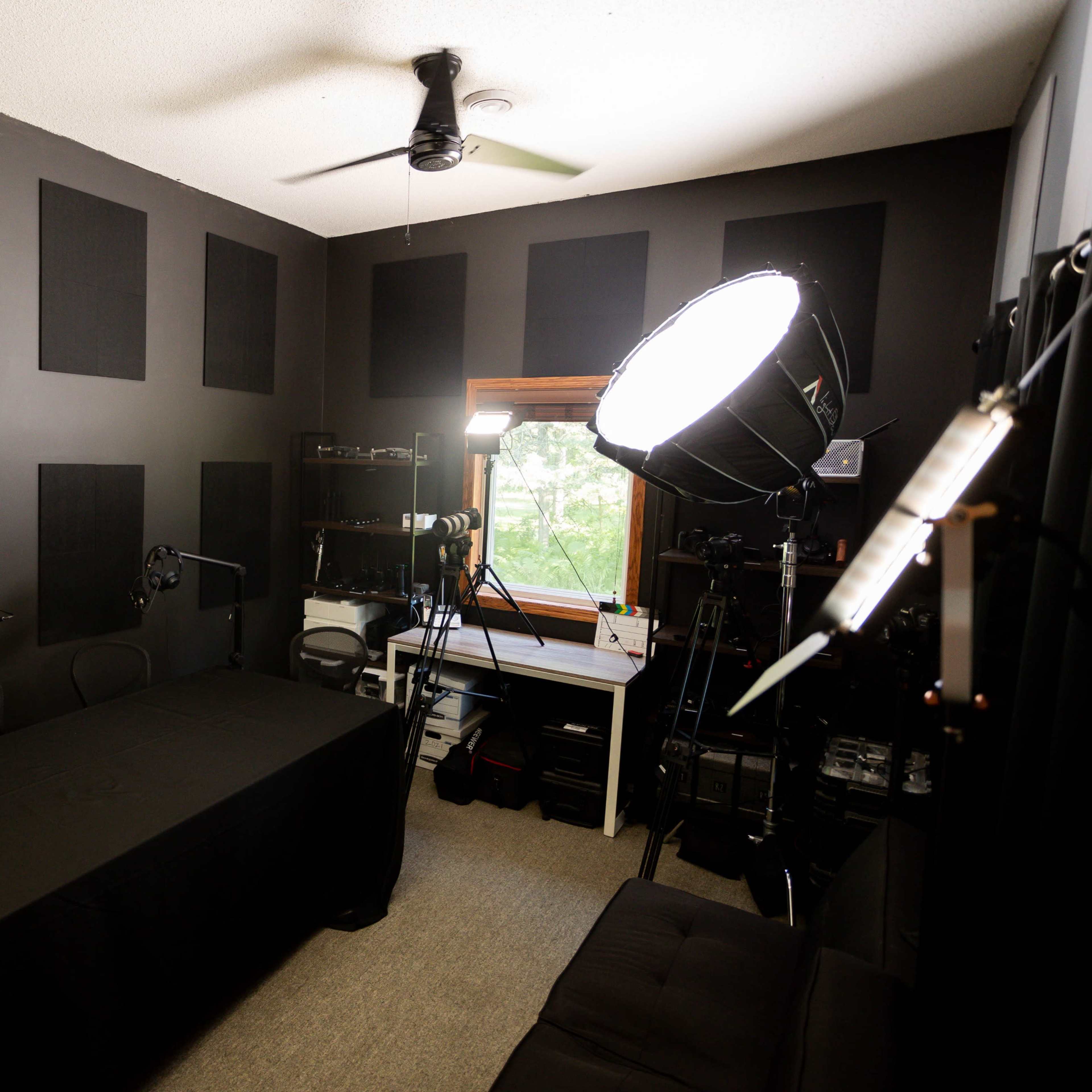 A recording studio with a dark panelled walls, a large table covered with a black cloth, several microphones on stands, and equipment positioned around the room, including a large light source and a window.