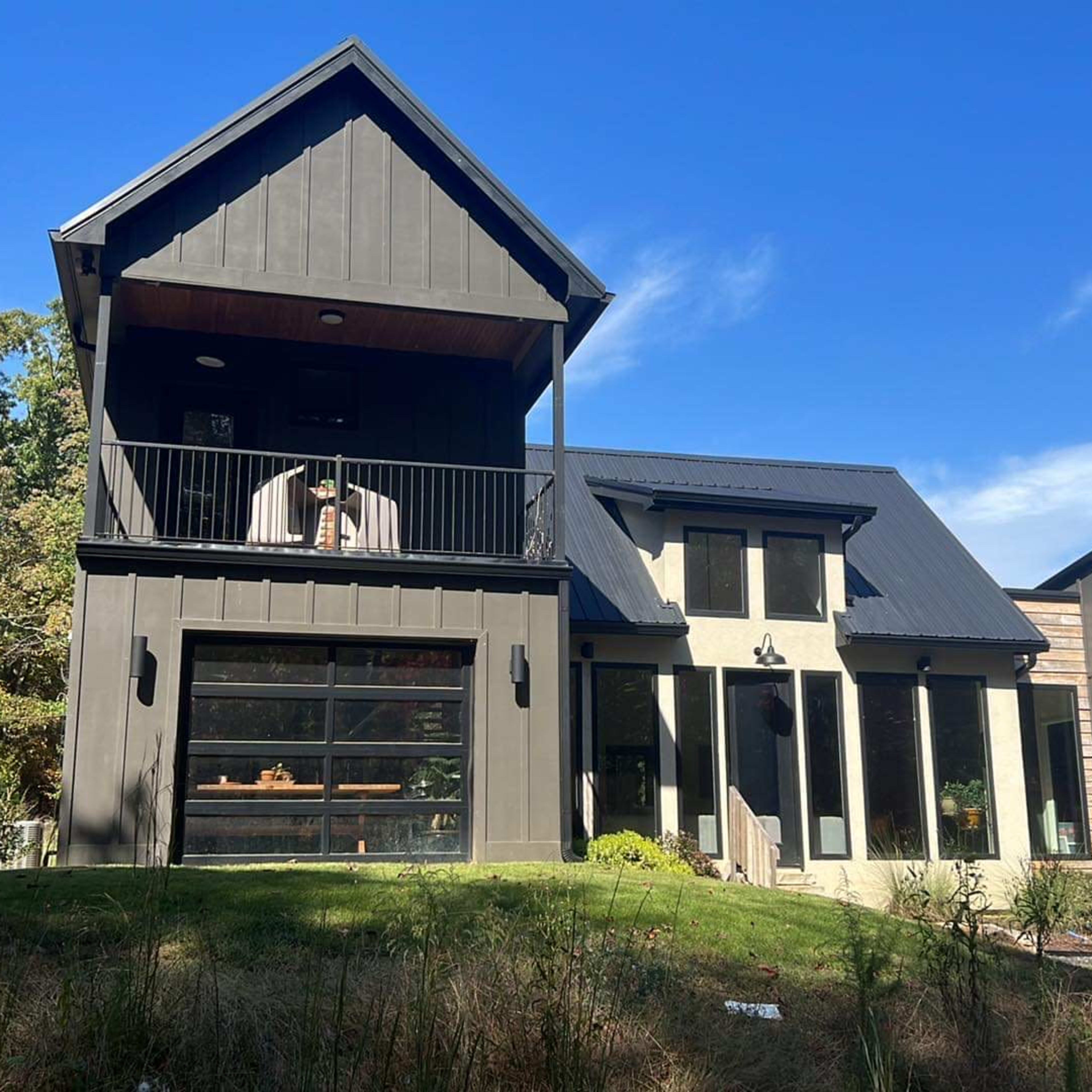 A modern black-clad house features a prominent balcony and large windows, surrounded by greenery and trees.