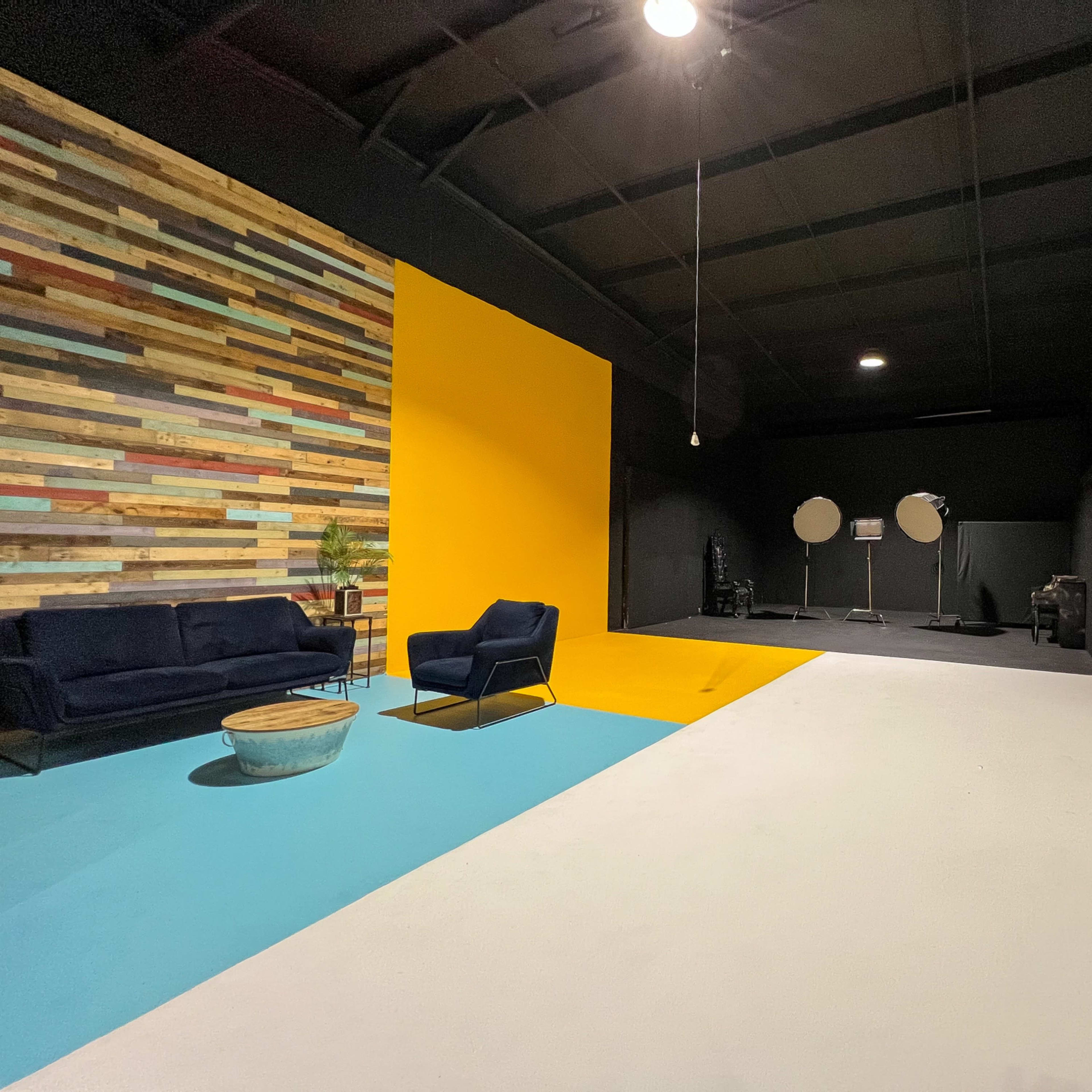 A spacious studio with a textured wooden accent wall, a bright yellow section, a light blue area, and a white backdrop, featuring a sofa and standing equipment.