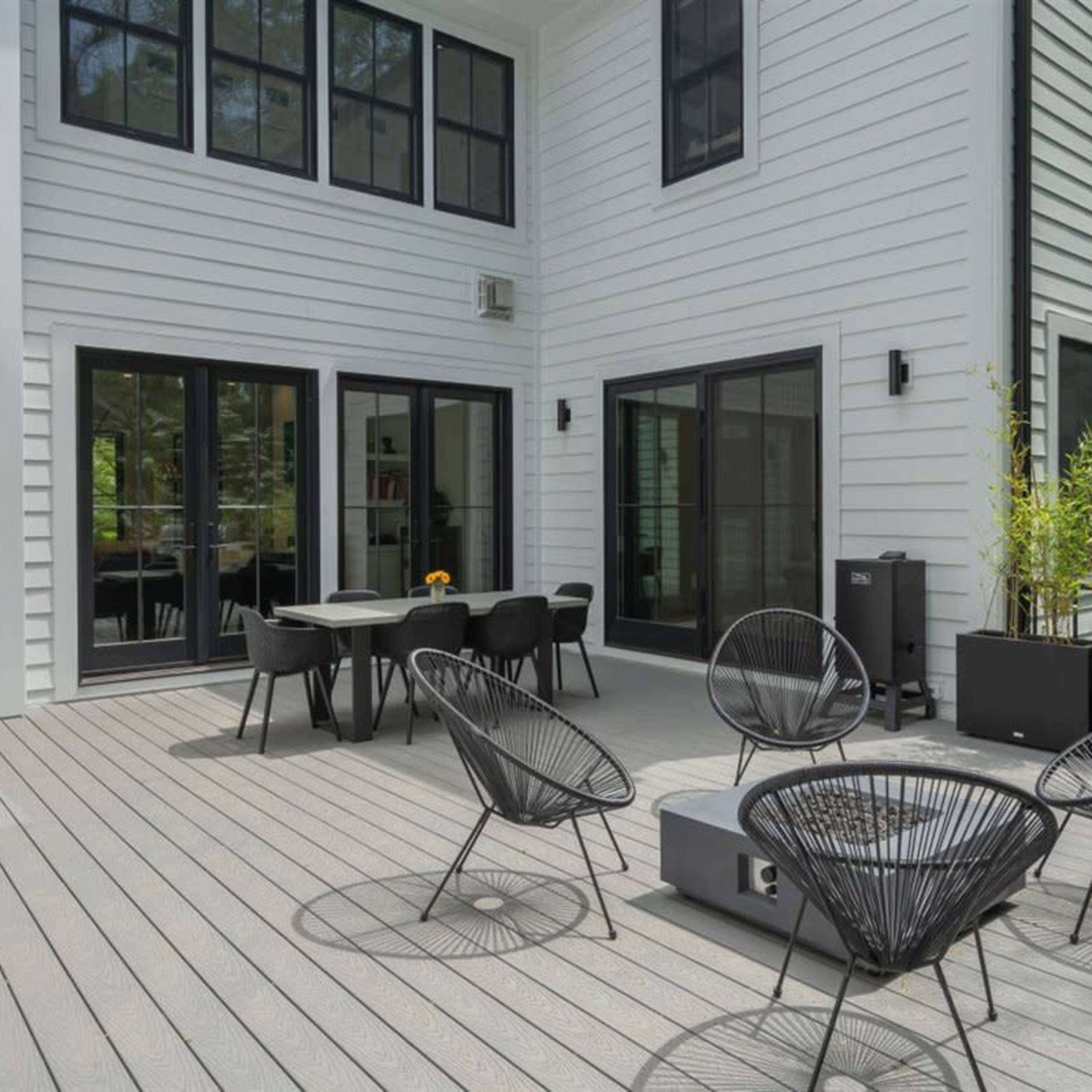 A modern outdoor patio area featuring black chairs, a table, and a grill against the backdrop of a white, two-story house surrounded by greenery.