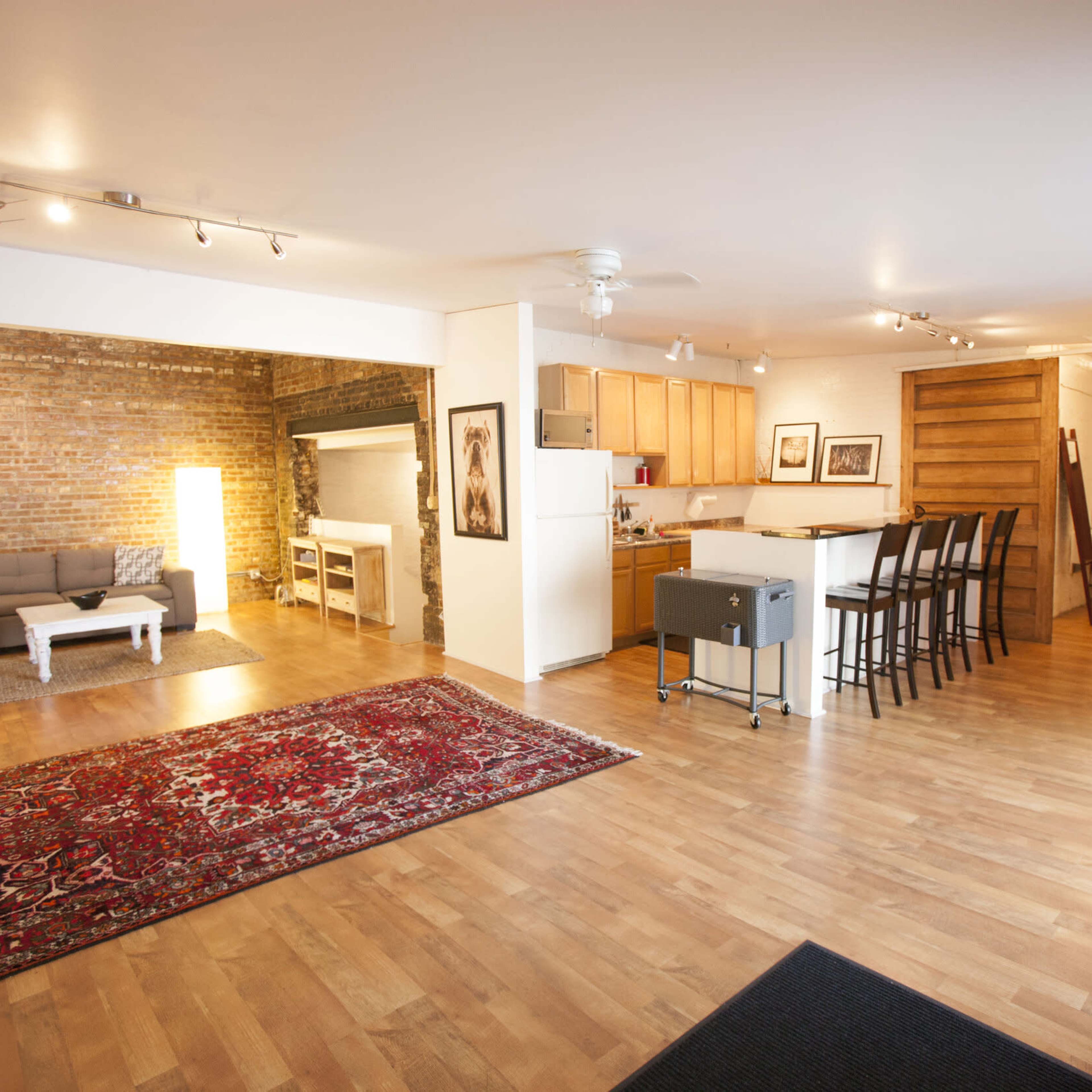 A spacious living area with hardwood floors, a large area rug, a sectional sofa, and a kitchen with wooden cabinets in an open-concept design.