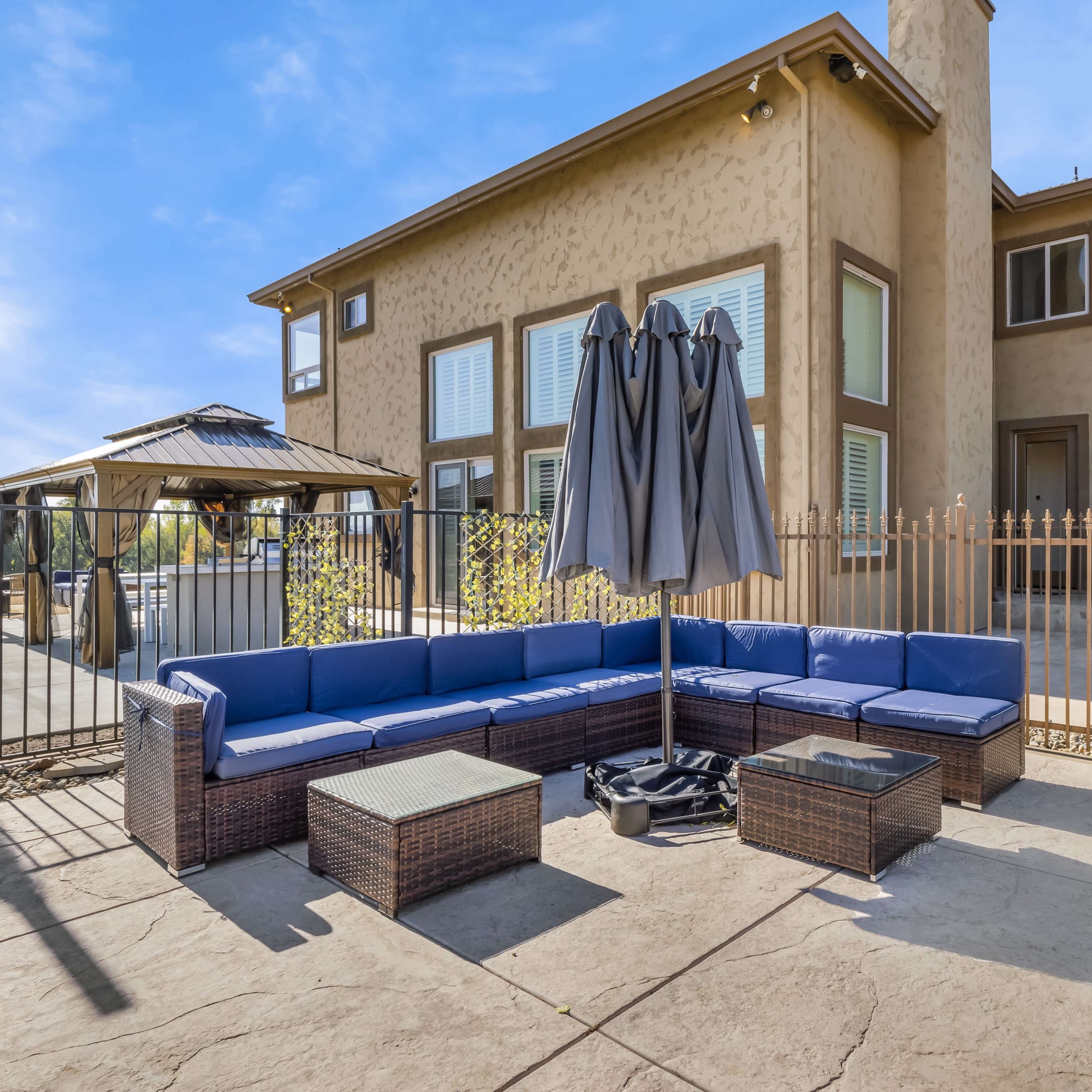A patio area features a blue sectional sofa arranged around a glass coffee table, alongside a large umbrella and a fenced yard with a building in the background.