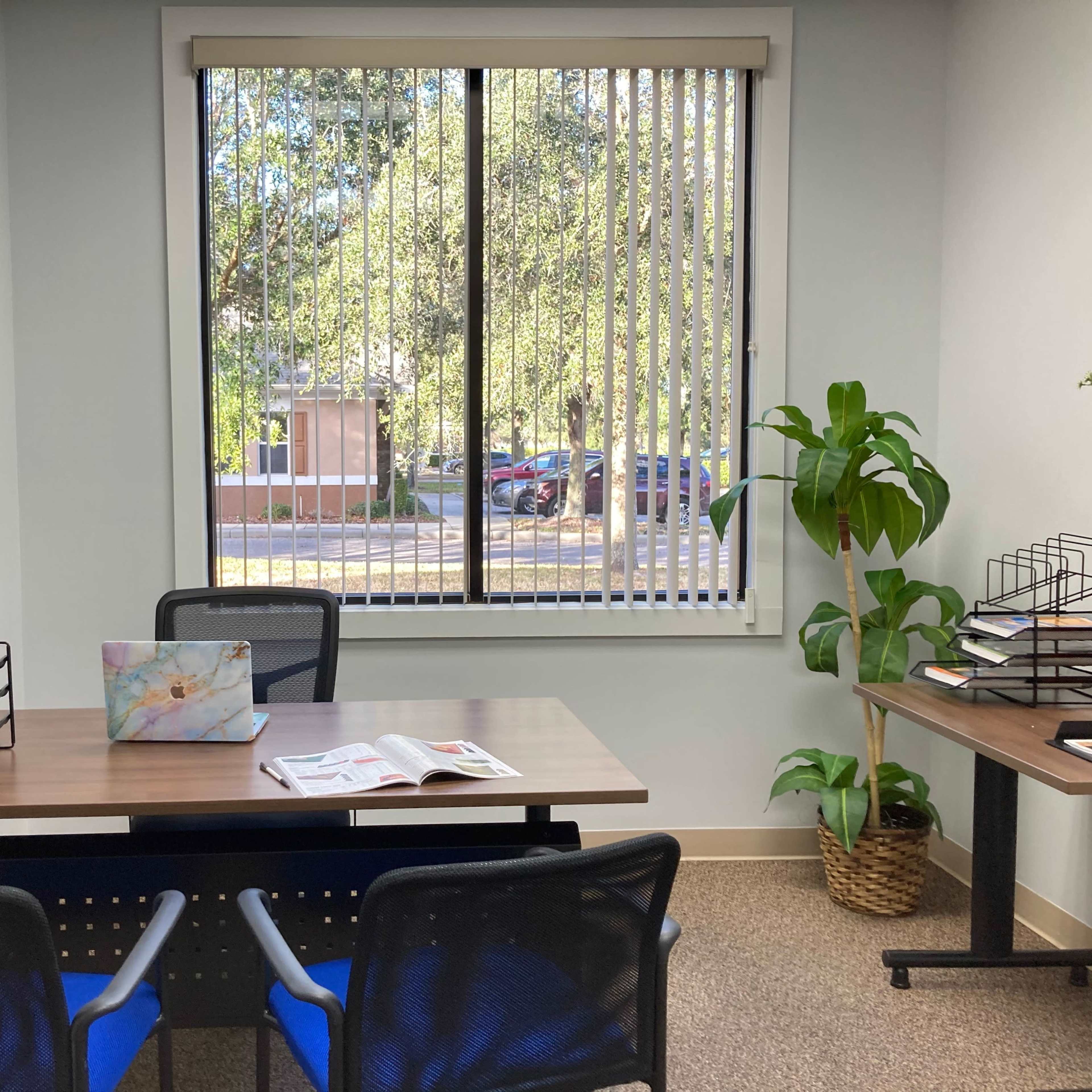 A small office with two empty desks, a plant, and a large window that overlooks a parking lot and trees.