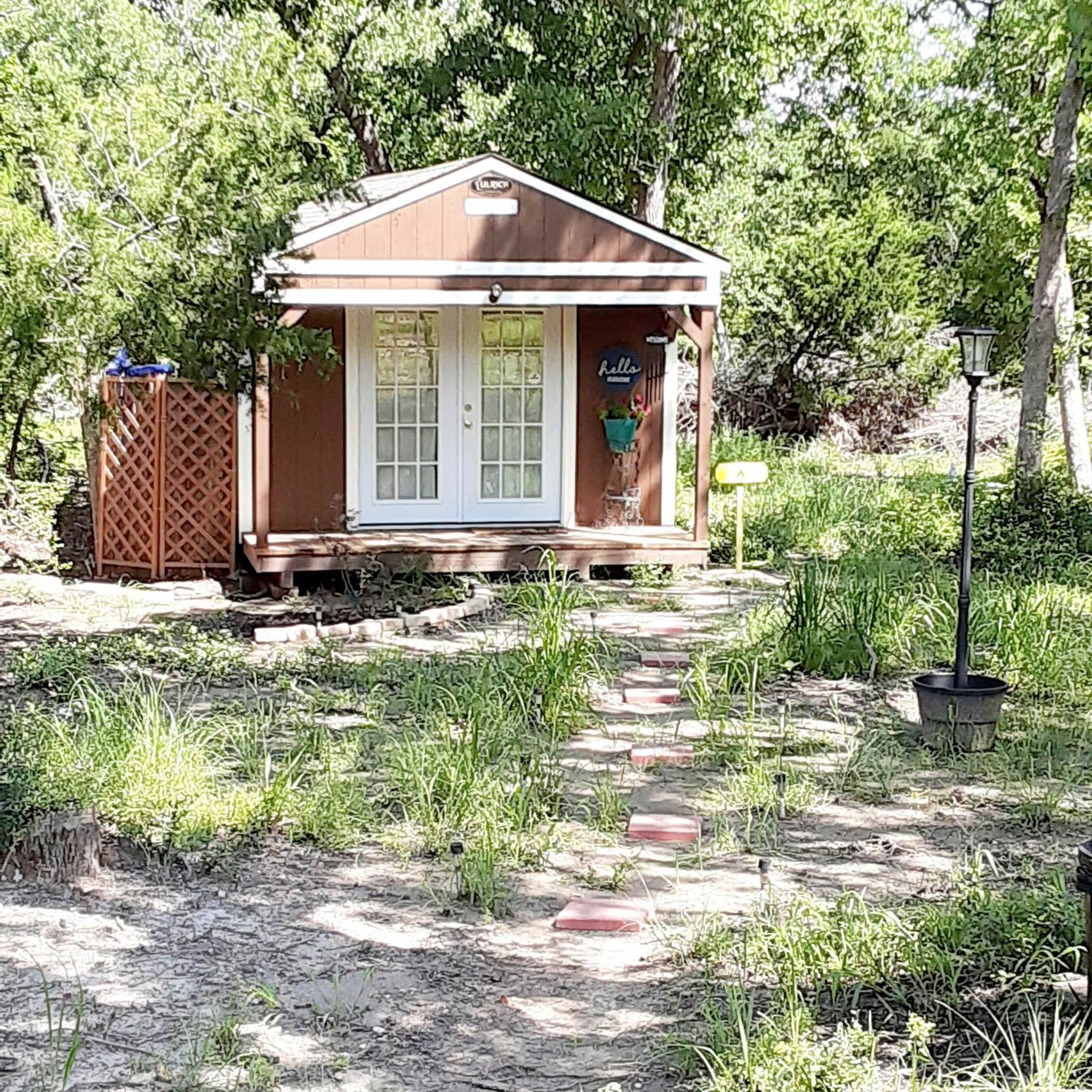 A small wooden cabin with a porch sits at the end of a stone path surrounded by grass and trees.