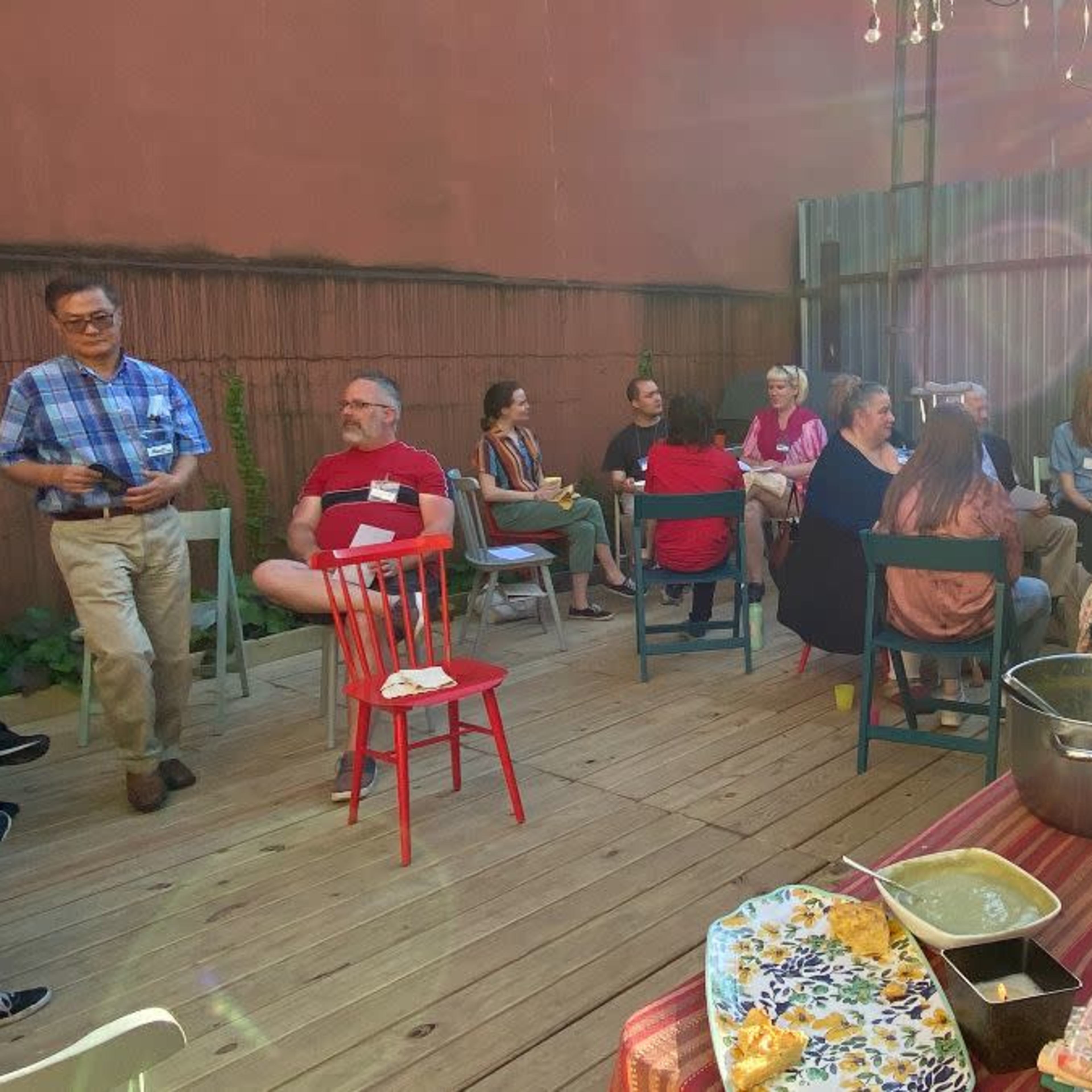 A group of people is seated in a courtyard, engaged in conversation, with several tables displaying food and drinks.