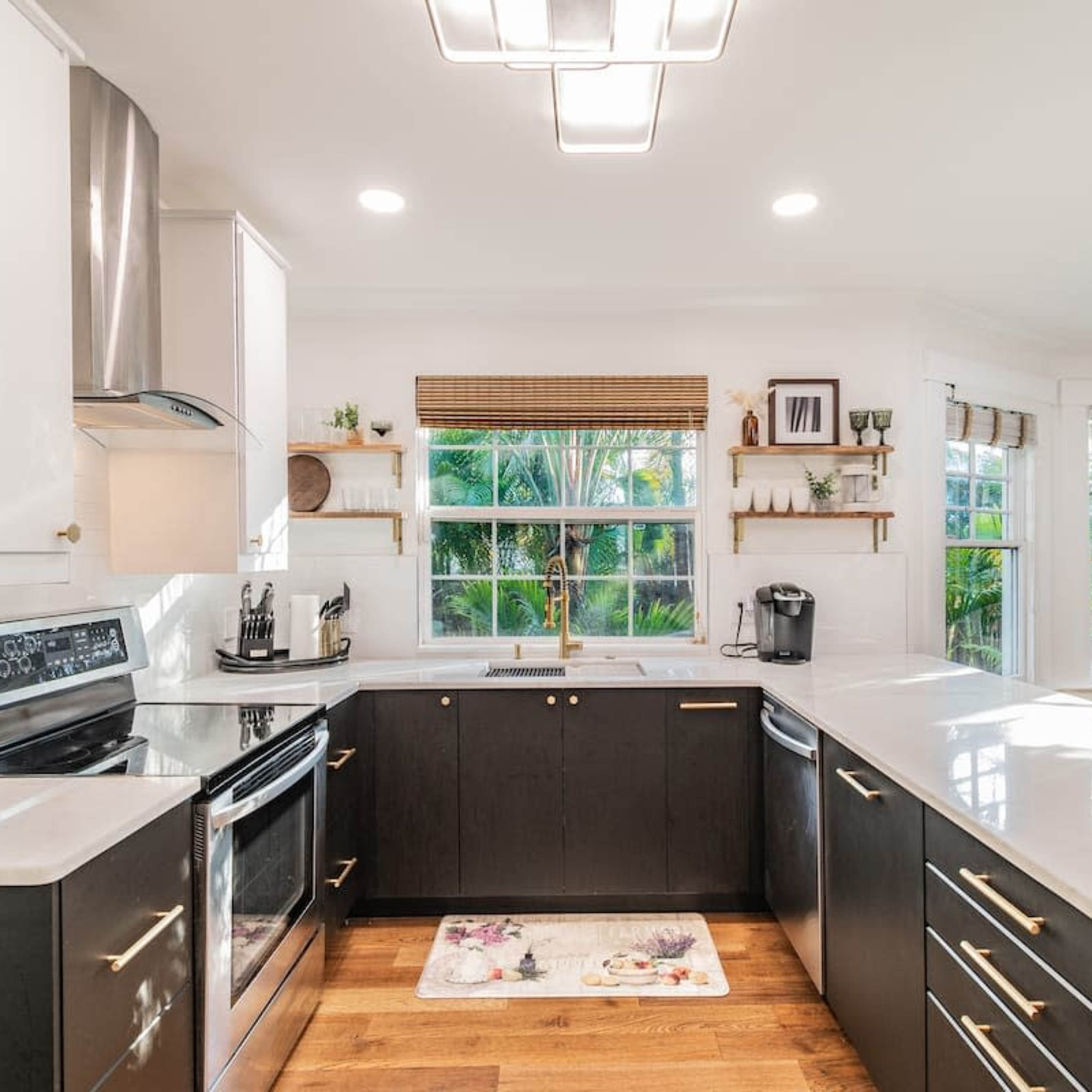 A modern kitchen features dark cabinets, stainless steel appliances, and a large island with a white countertop, surrounded by windows that offer a view of greenery outside.