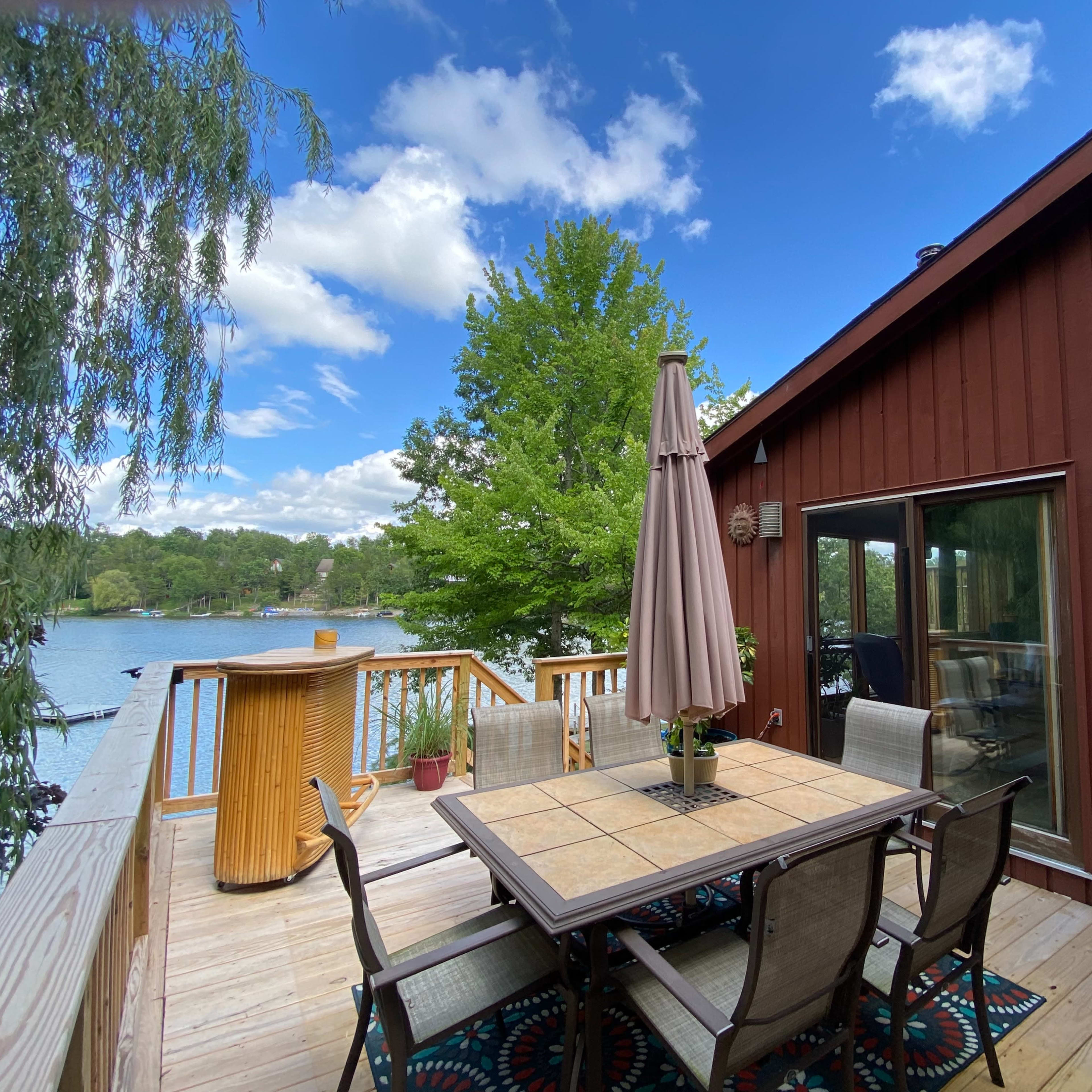 A wooden deck with a table and chairs is set up by a lake, surrounded by greenery and under a partly cloudy sky.