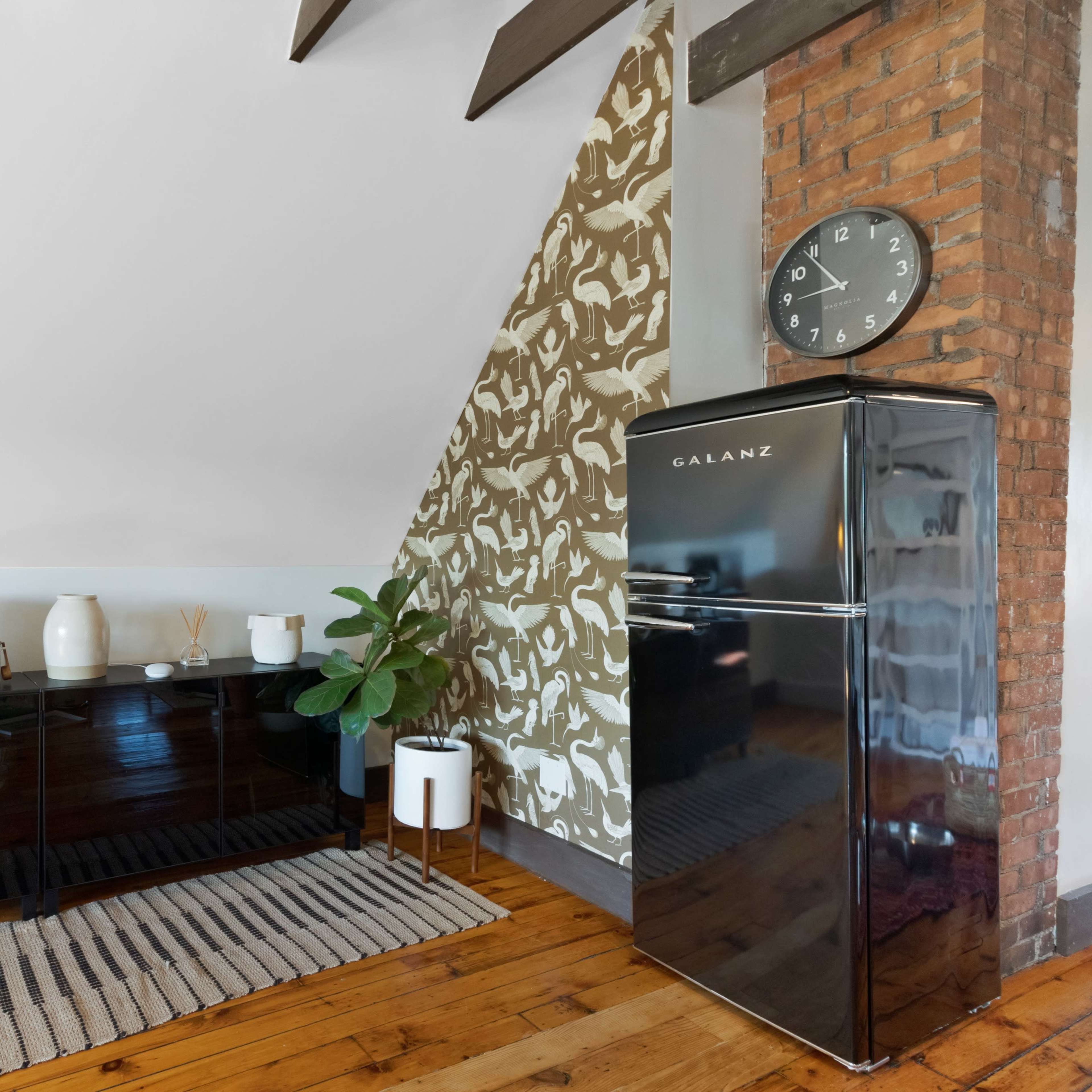A corner of a kitchen featuring a black refrigerator, a dark cabinet, a plant, and a clock on a brick wall, with patterned wallpaper and wooden flooring.