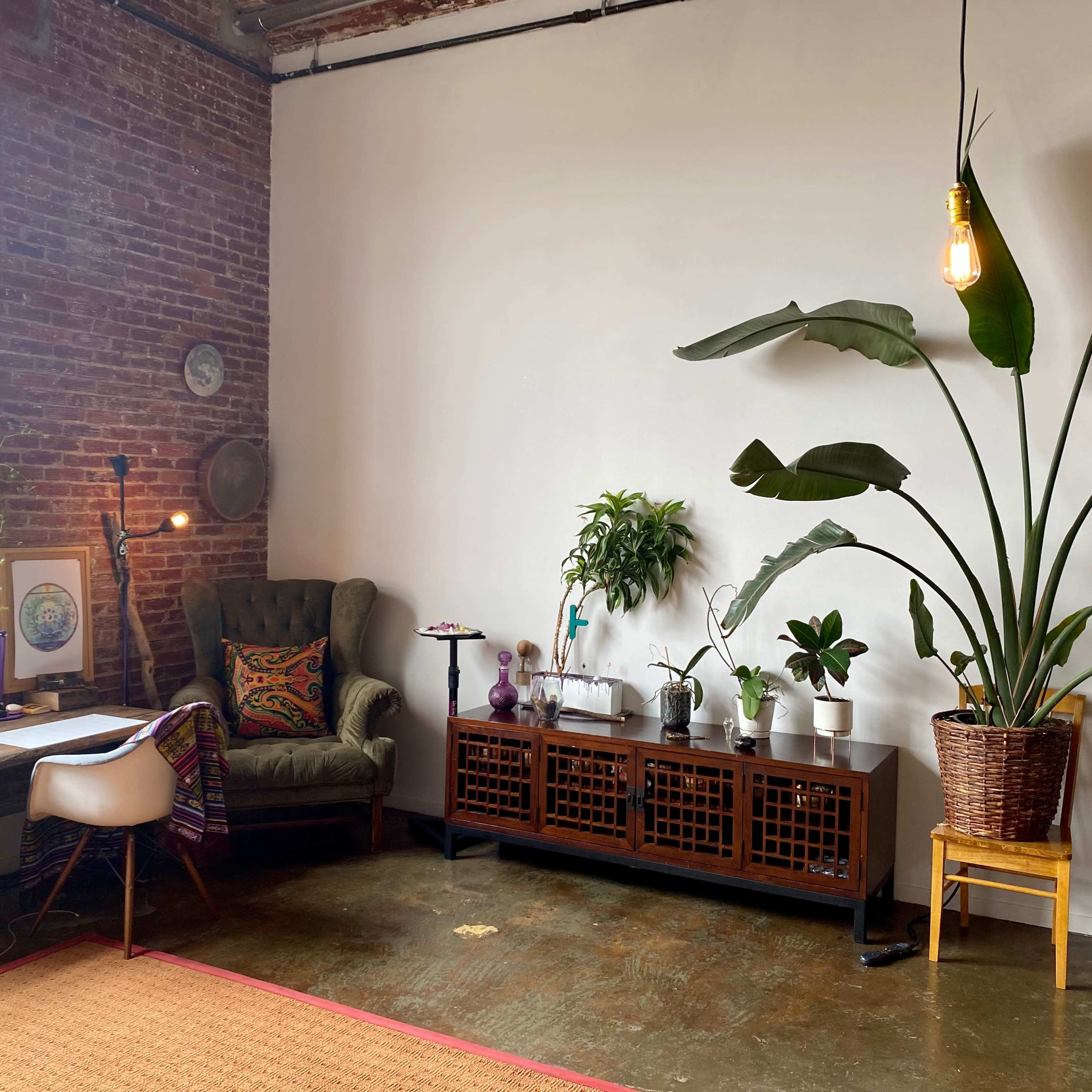 A cozy room with a brown brick wall, a wooden desk and chair, a large potted plant, and a cabinet decorated with various indoor plants.