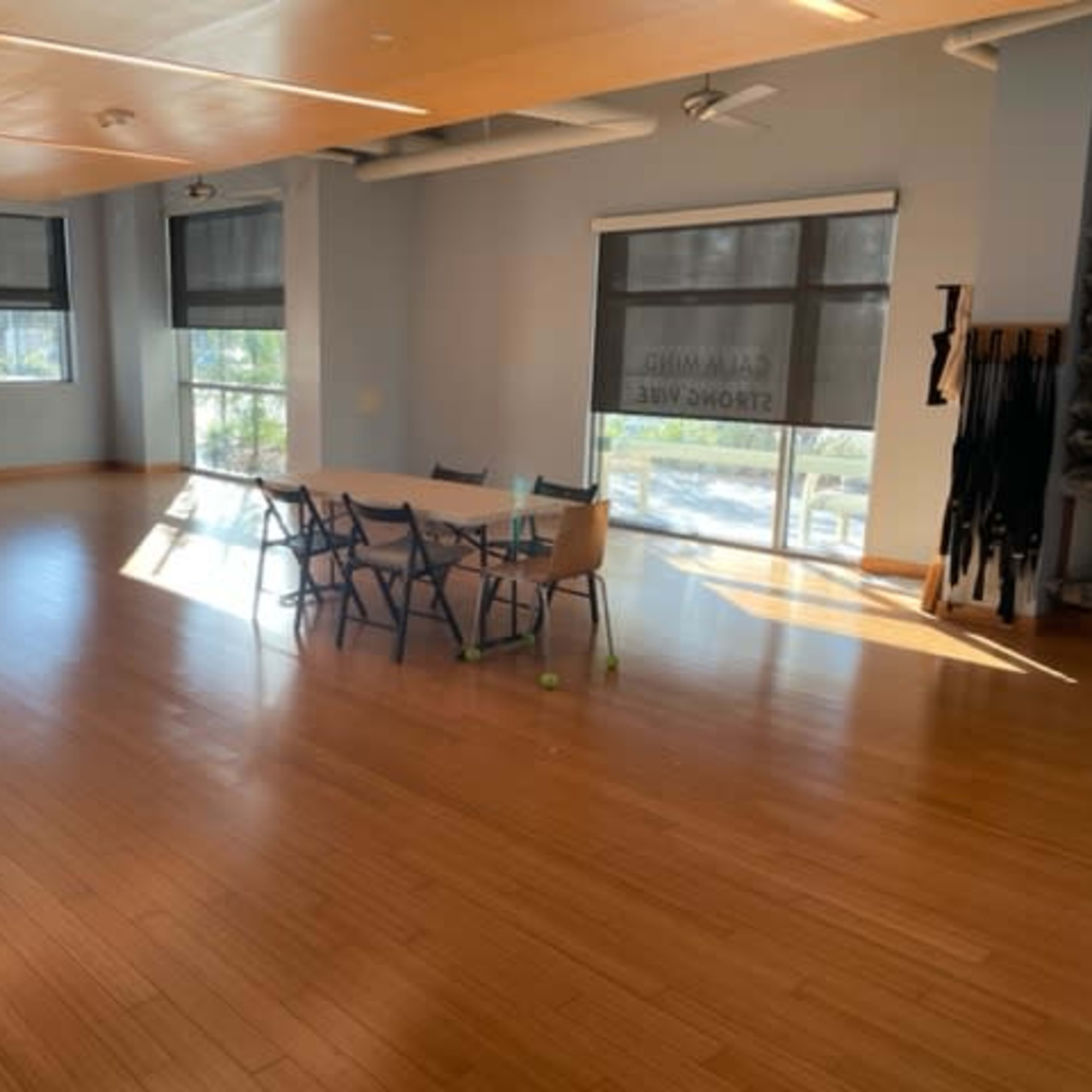 A spacious room with wooden flooring features a central table surrounded by chairs, large windows with blinds, and minimal furnishings along the walls.