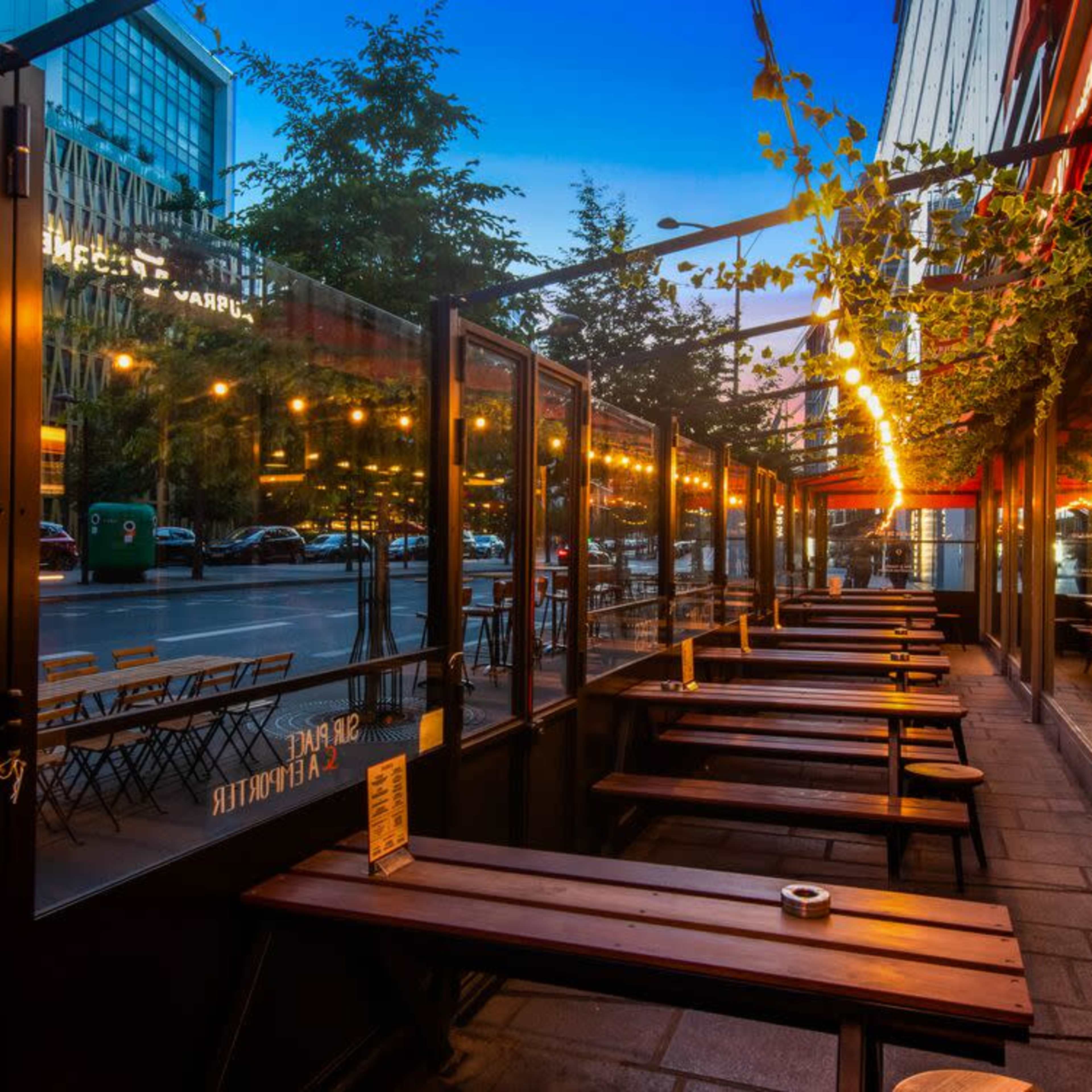 An outdoor dining area with wooden tables and benches, lined by glass panels, illuminated by string lights, and facing a street in an urban setting at dusk.