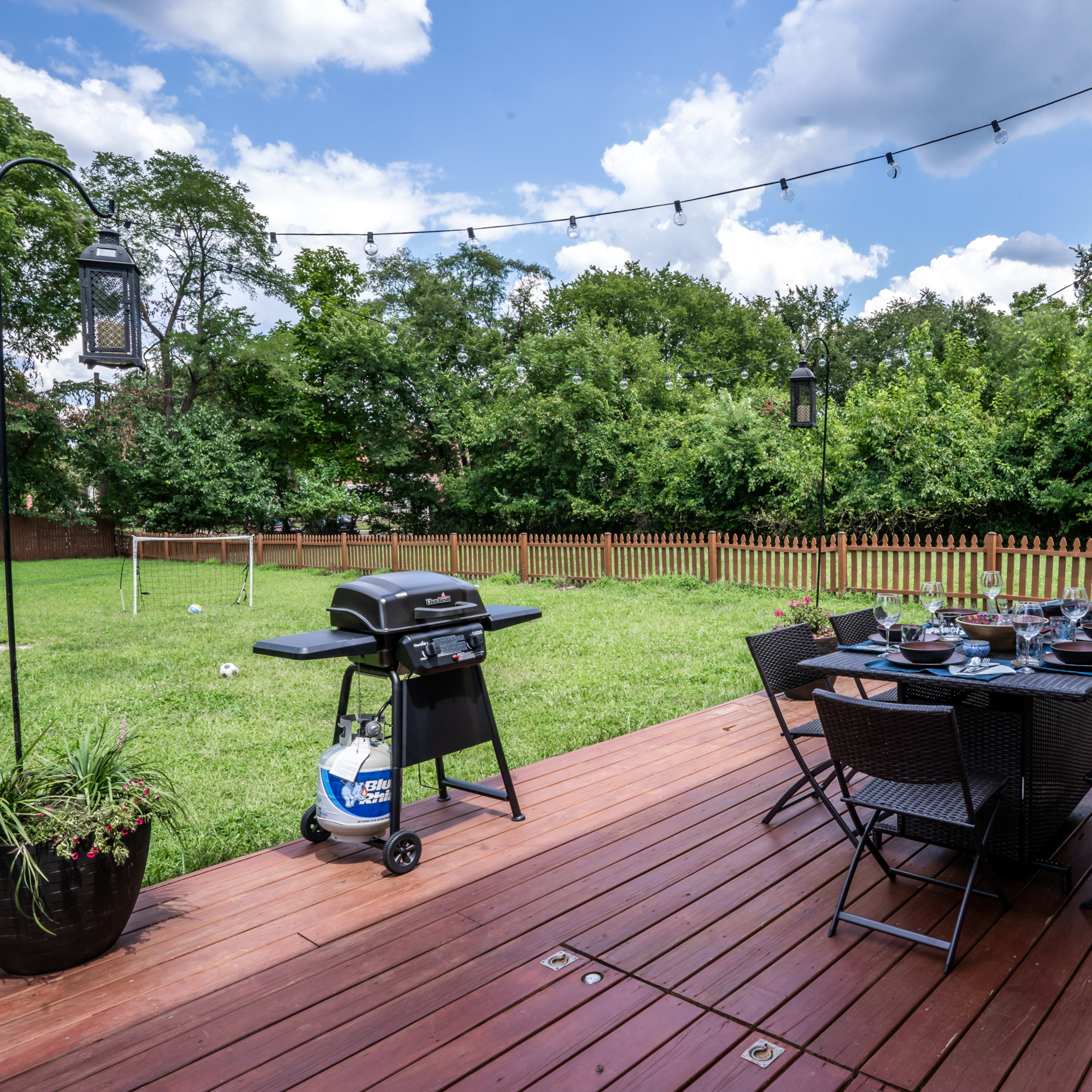A wooden deck features a dining table set with dishes and a grill, surrounded by a green lawn and trees in the background.
