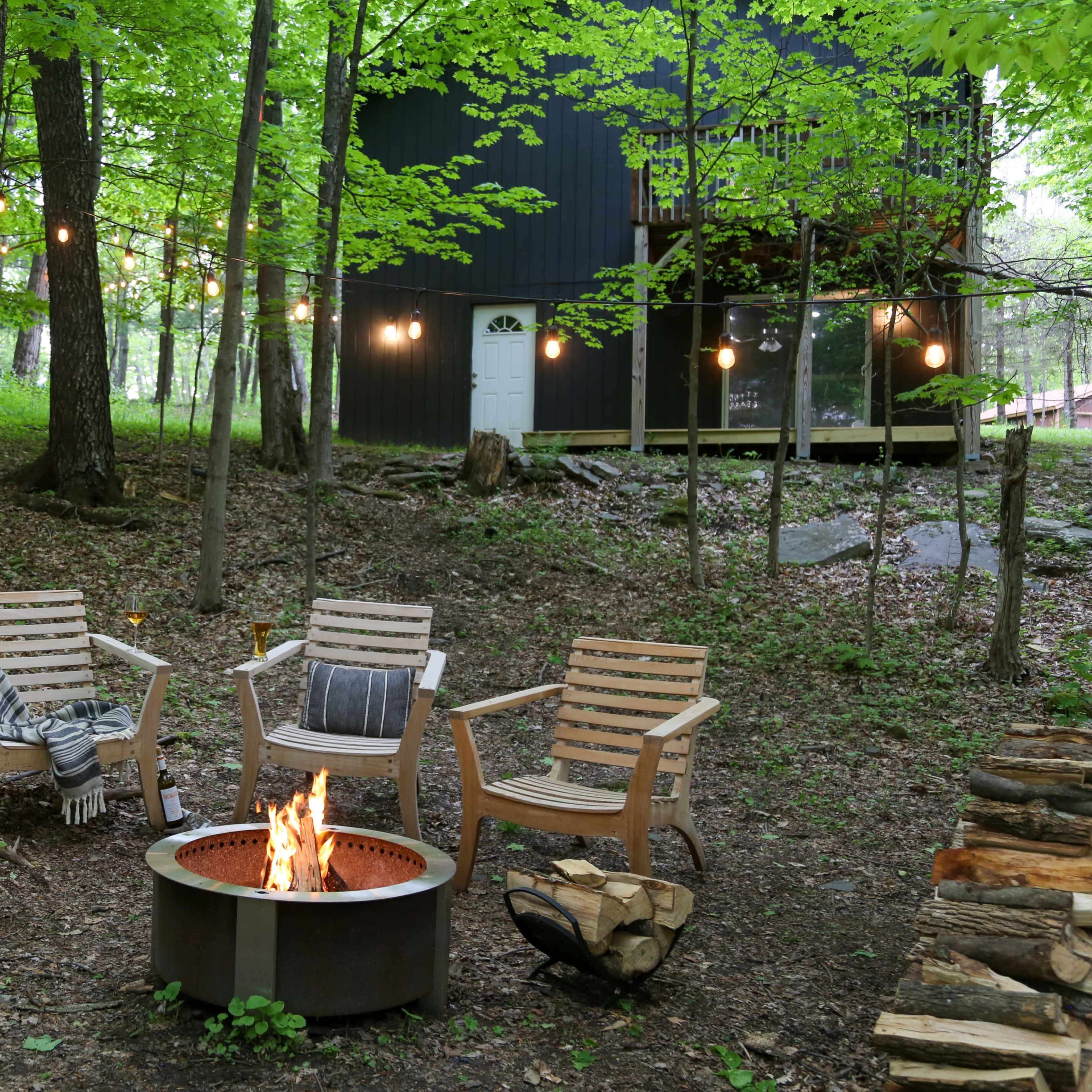 A rustic fire pit surrounded by wooden chairs is situated in a wooded area, with string lights hanging above and a black cabin in the background.