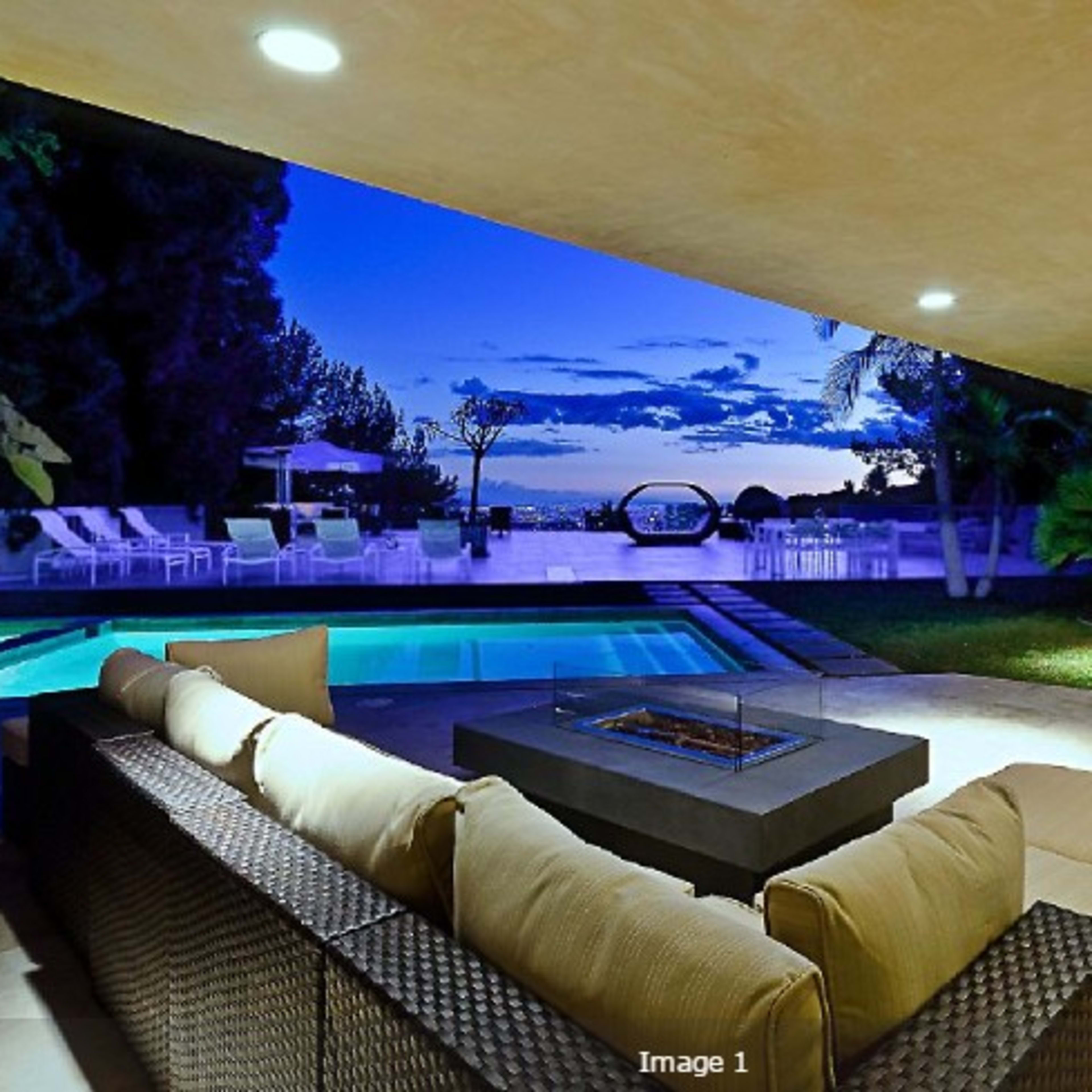 A modern outdoor living area features a sectional sofa with a fire pit, overlooking a swimming pool and a scenic twilight sky.