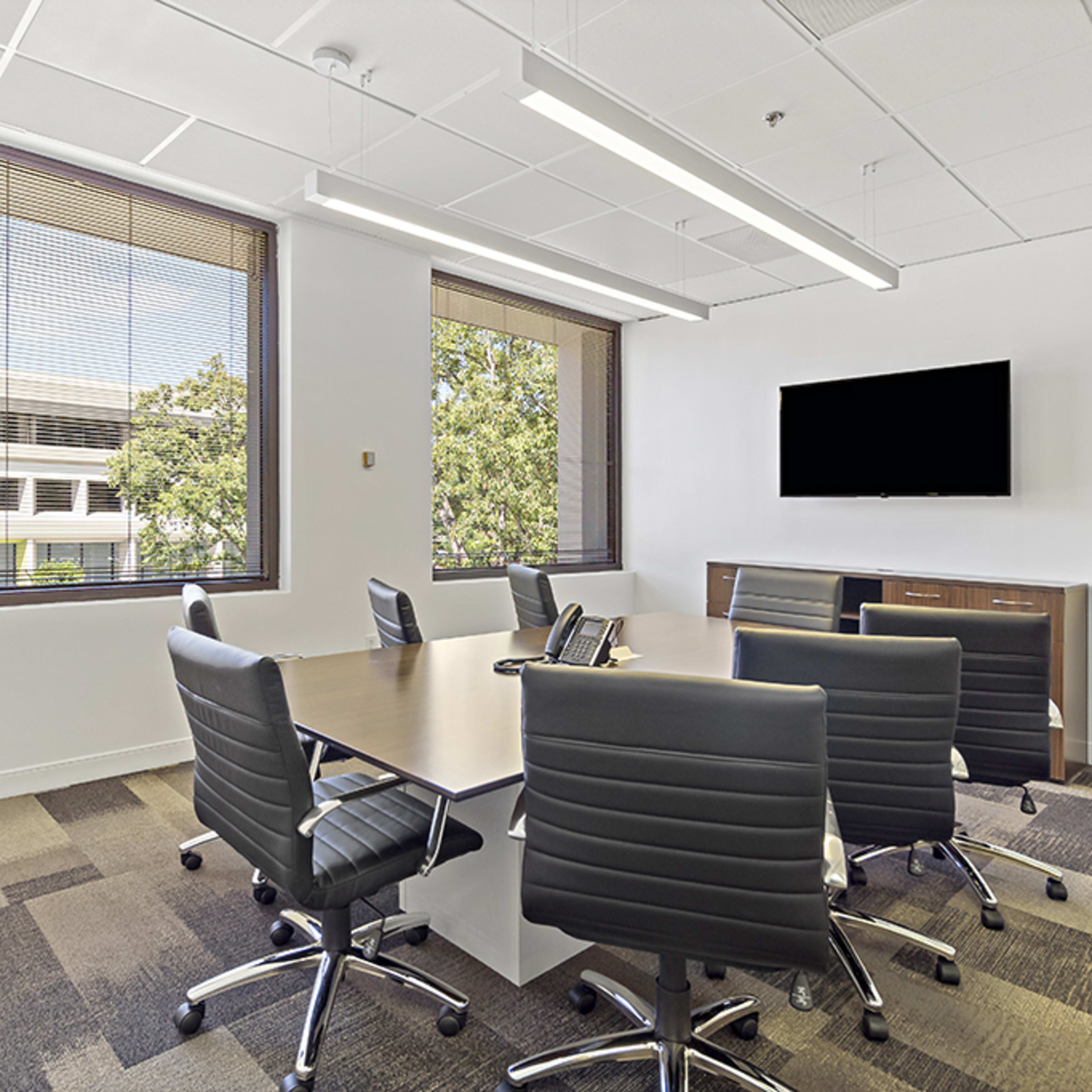 A conference room features a large table surrounded by black leather chairs, with windows offering a view of the outdoors and a wall-mounted television.