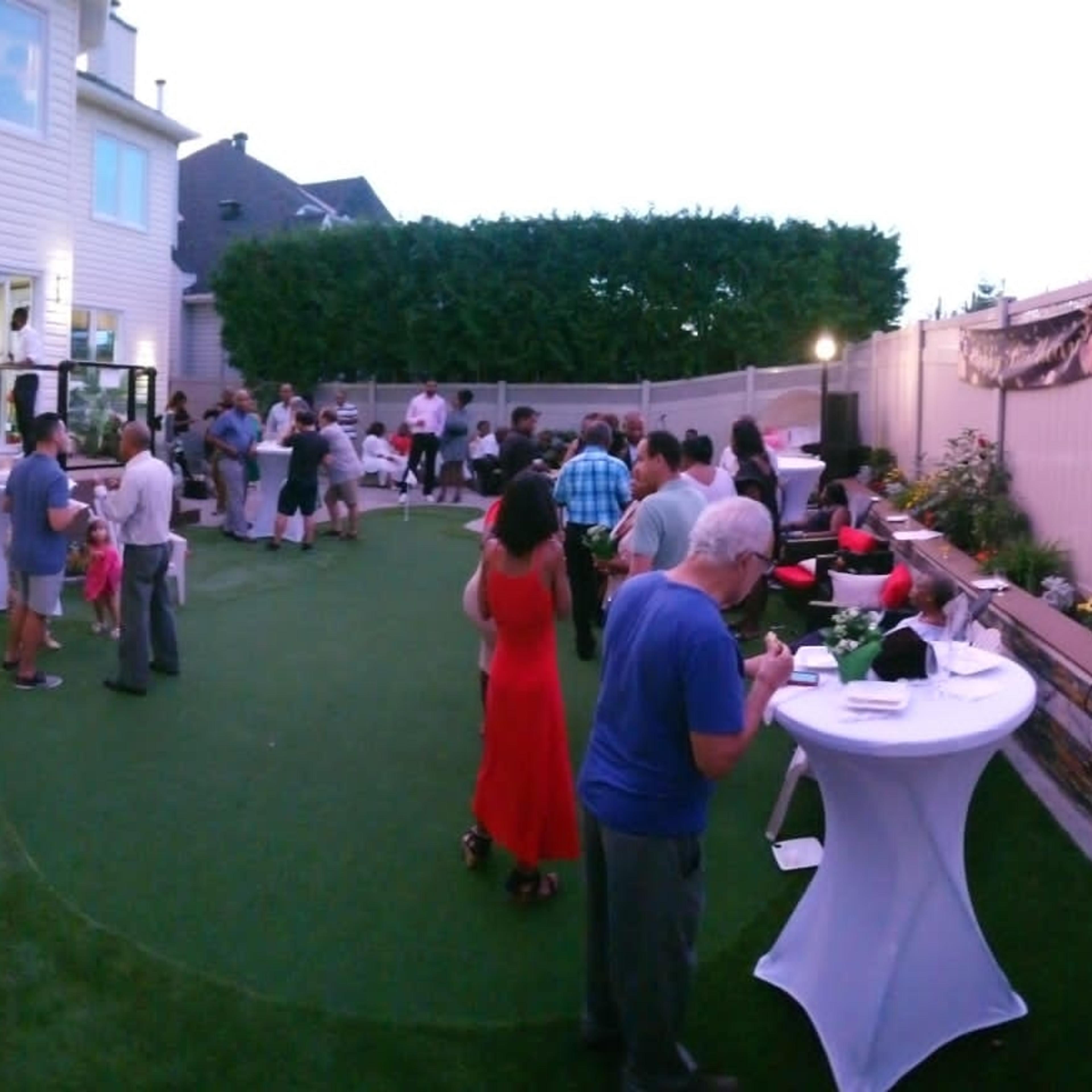 A gathering of people is taking place in a backyard with grass-like turf and a two-story house visible in the background.