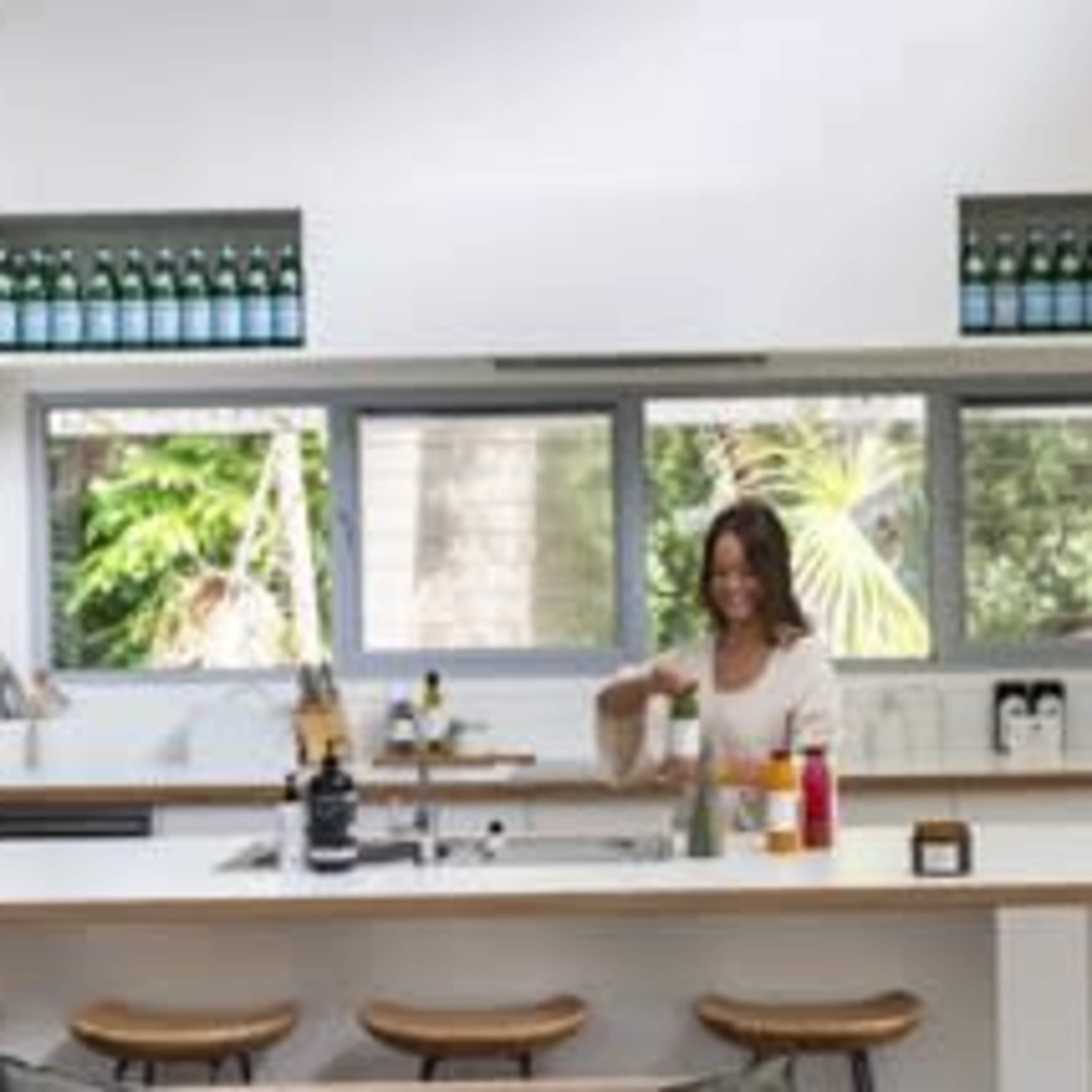 A woman prepares a drink at a modern kitchen island with bottles displayed on the shelves above and a view of greenery through large windows.