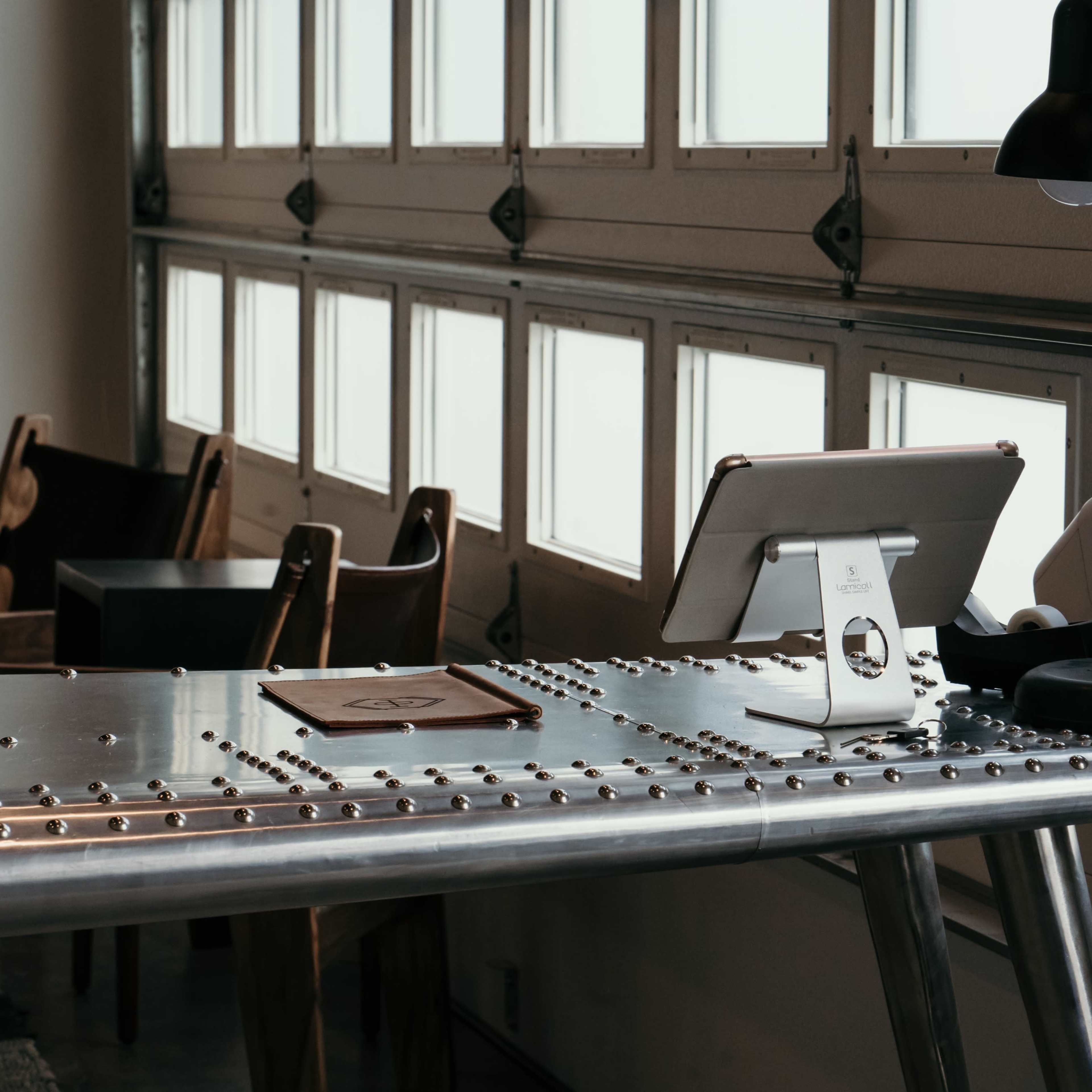 A metallic table with rivets and an iPad stand is situated in a room with large windows and wooden chairs.