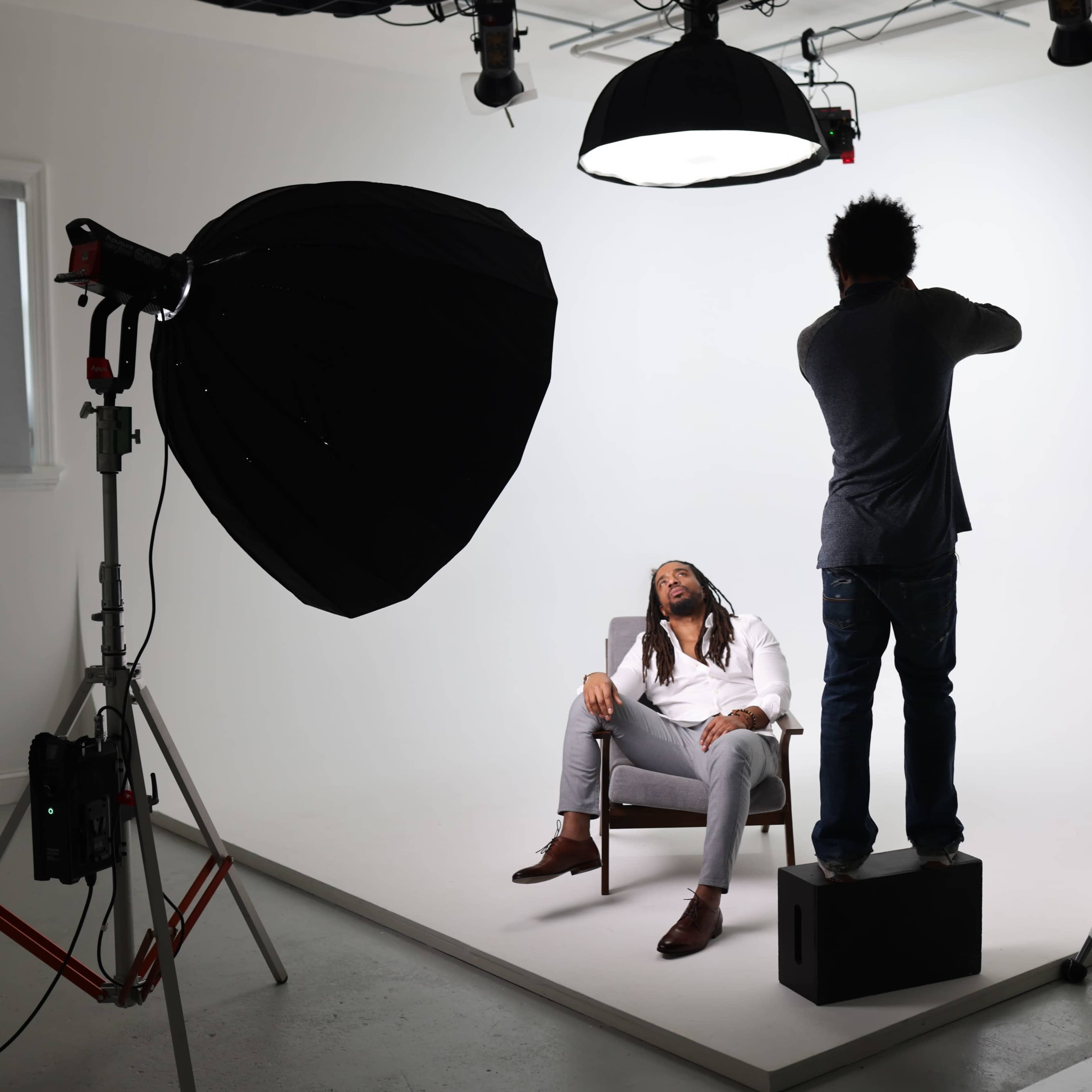 A photographer captures a seated model in a well-lit studio with white backdrops and professional lighting equipment.