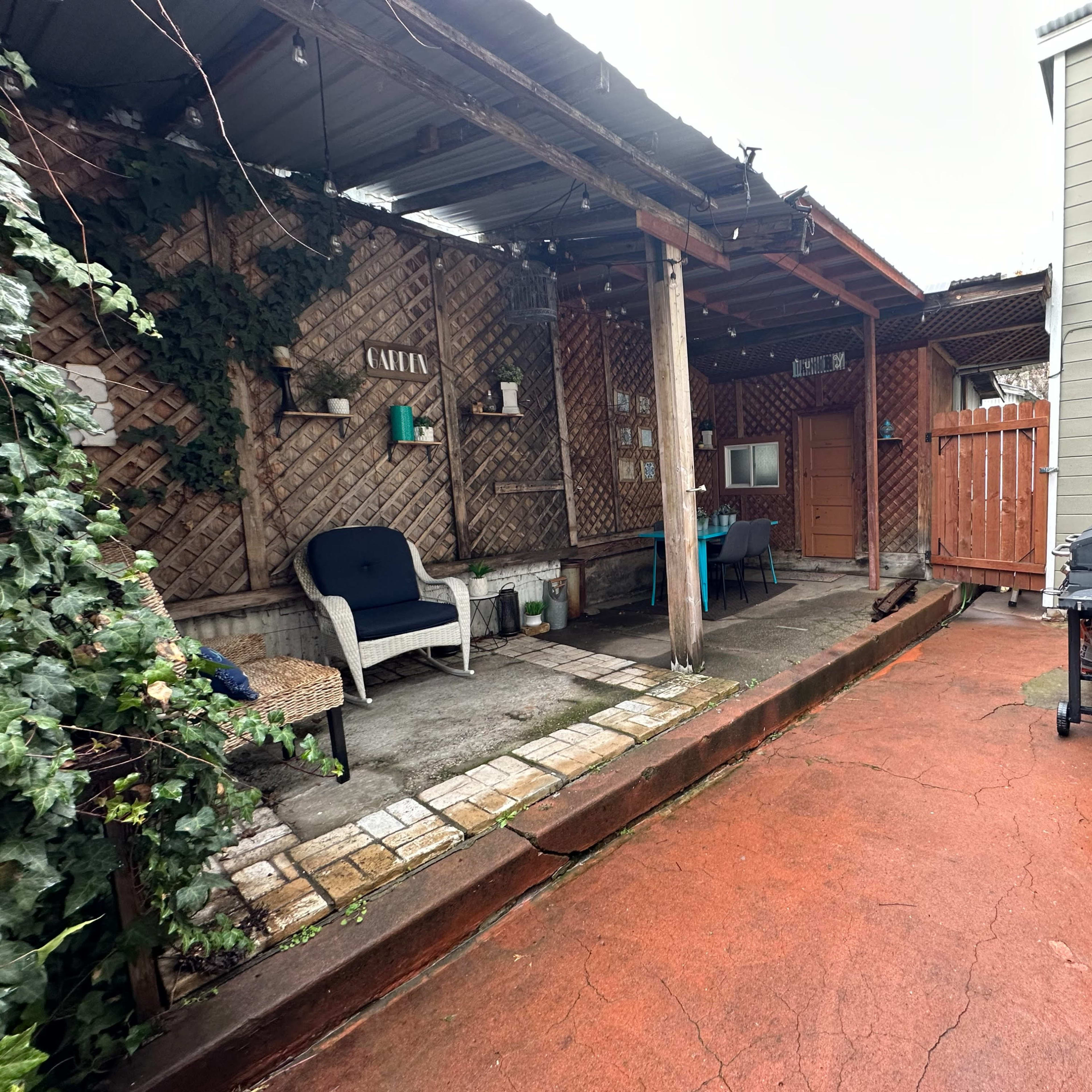 A narrow outdoor patio area features a gravel surface, wooden posts, a small table, and a grill, surrounded by walls and foliage.