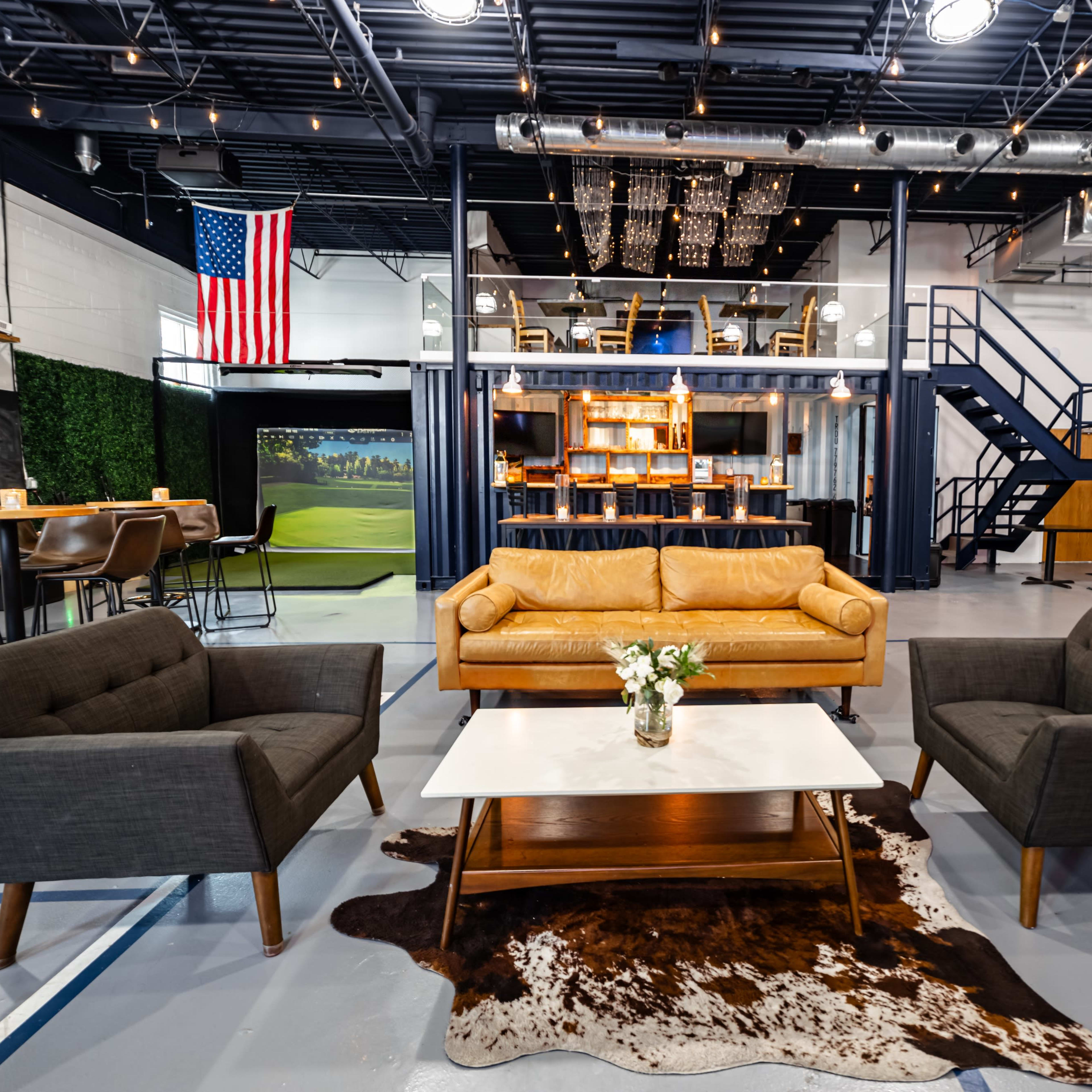 A spacious modern lounge area with a yellow leather couch, two gray armchairs, a coffee table on a cowhide rug, and a staircase leading to an upper level.