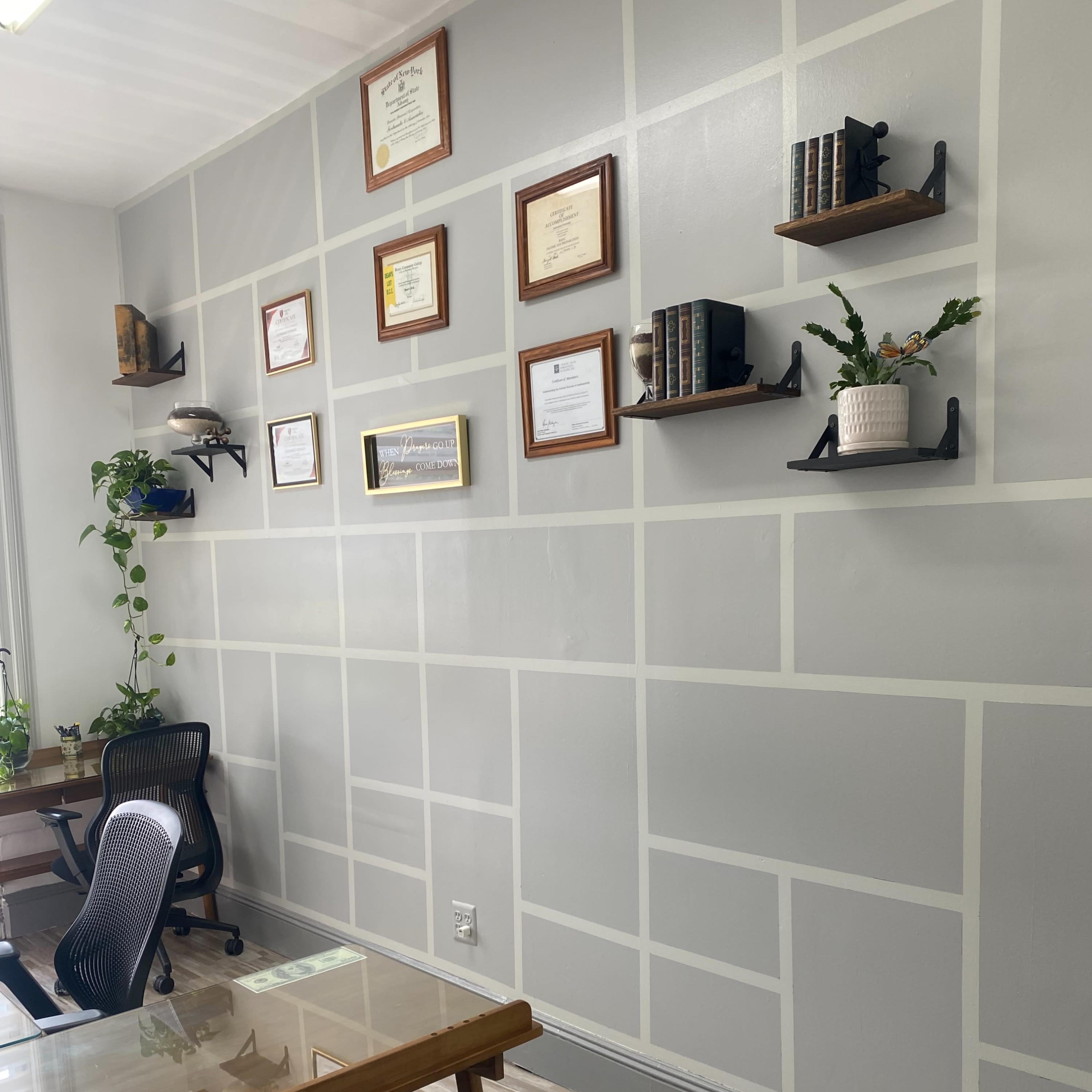 A modern office space with a wall featuring framed certificates and books on shelves, alongside a glass table and black chairs.