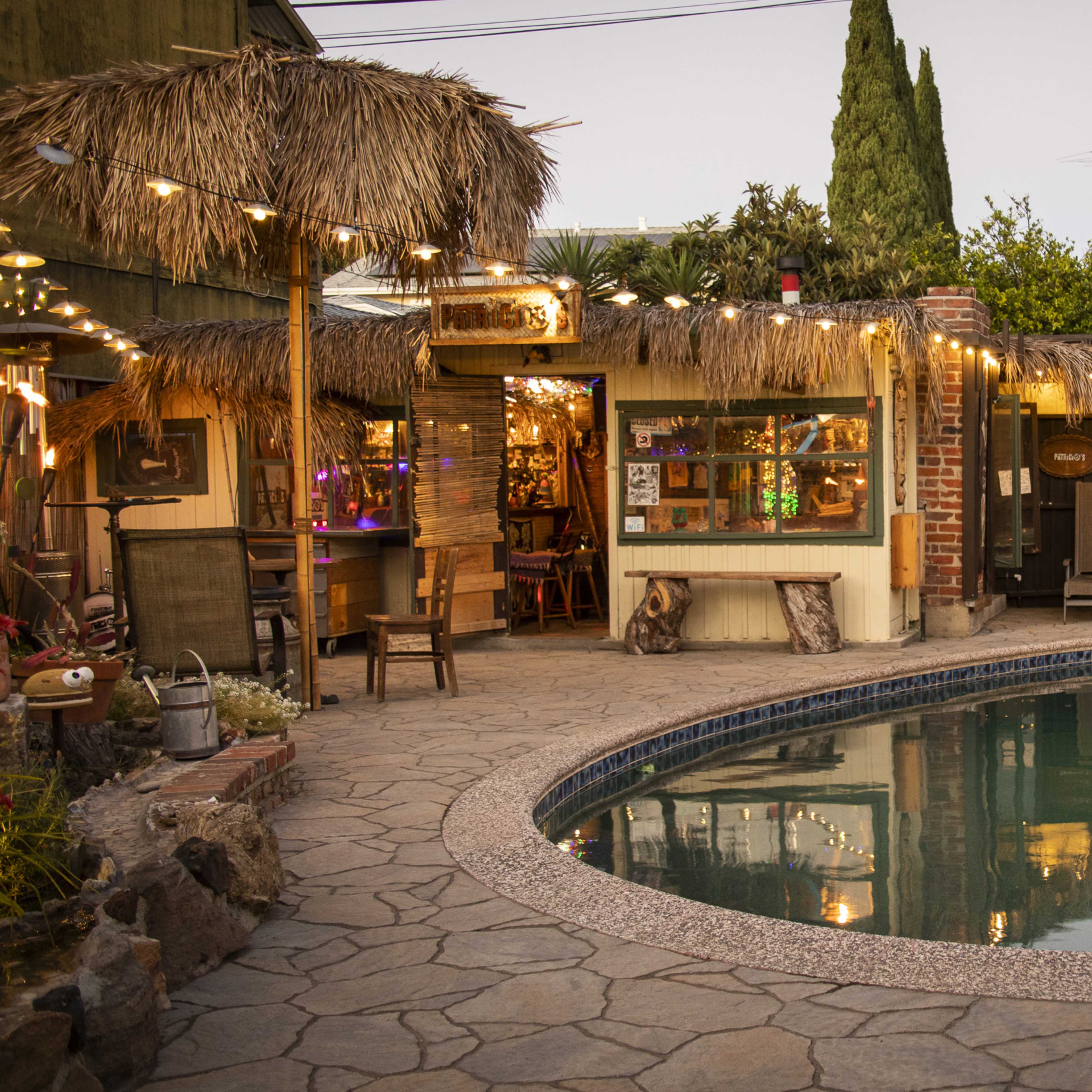 The scene features a backyard with a pool surrounded by stone paving, a thatched-roof bar area, and string lights overhead.