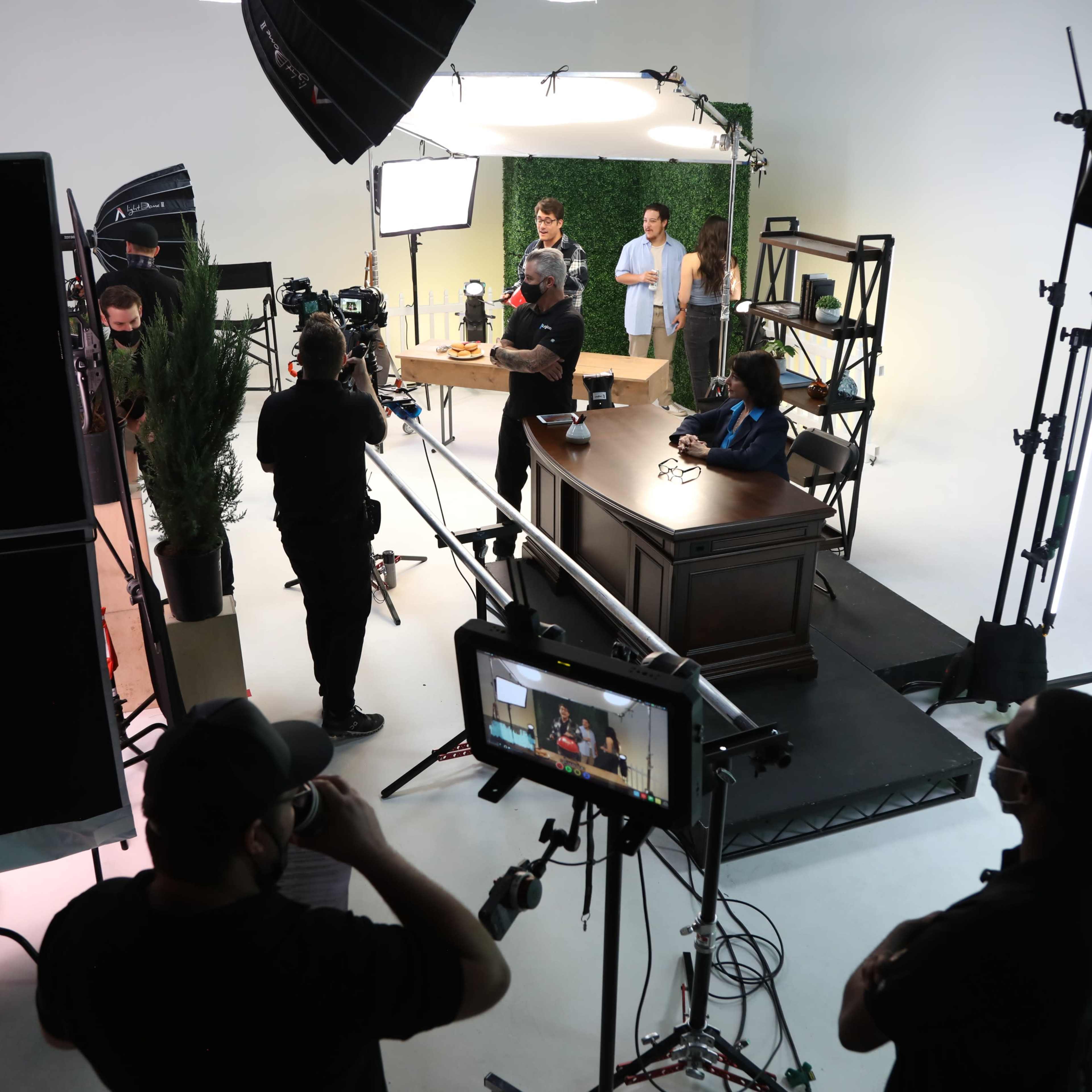 A film crew sets up equipment around a desk in a studio, with lights and cameras positioned for a production.