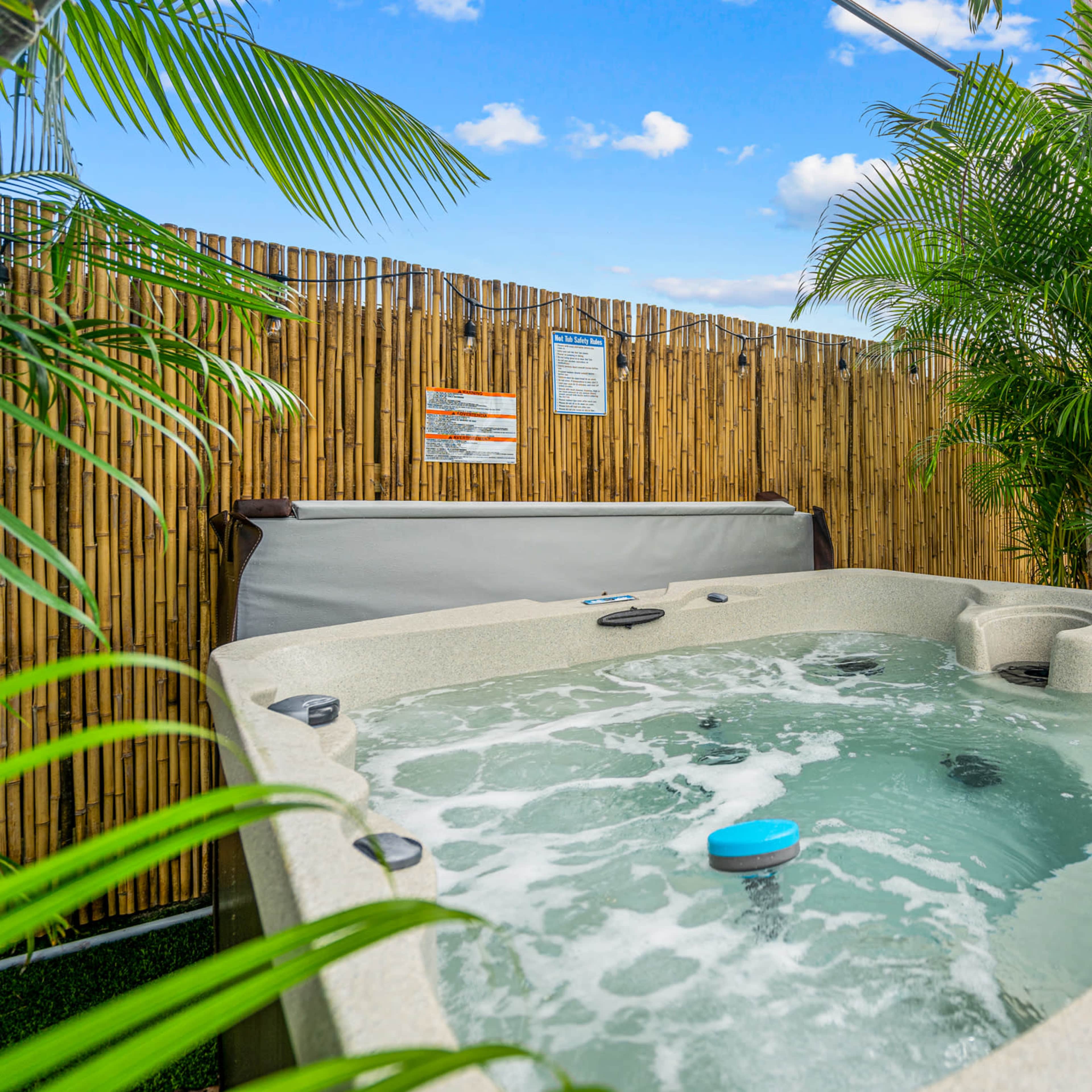A hot tub with bubbling water is surrounded by palm plants and a bamboo fence in a backyard setting.