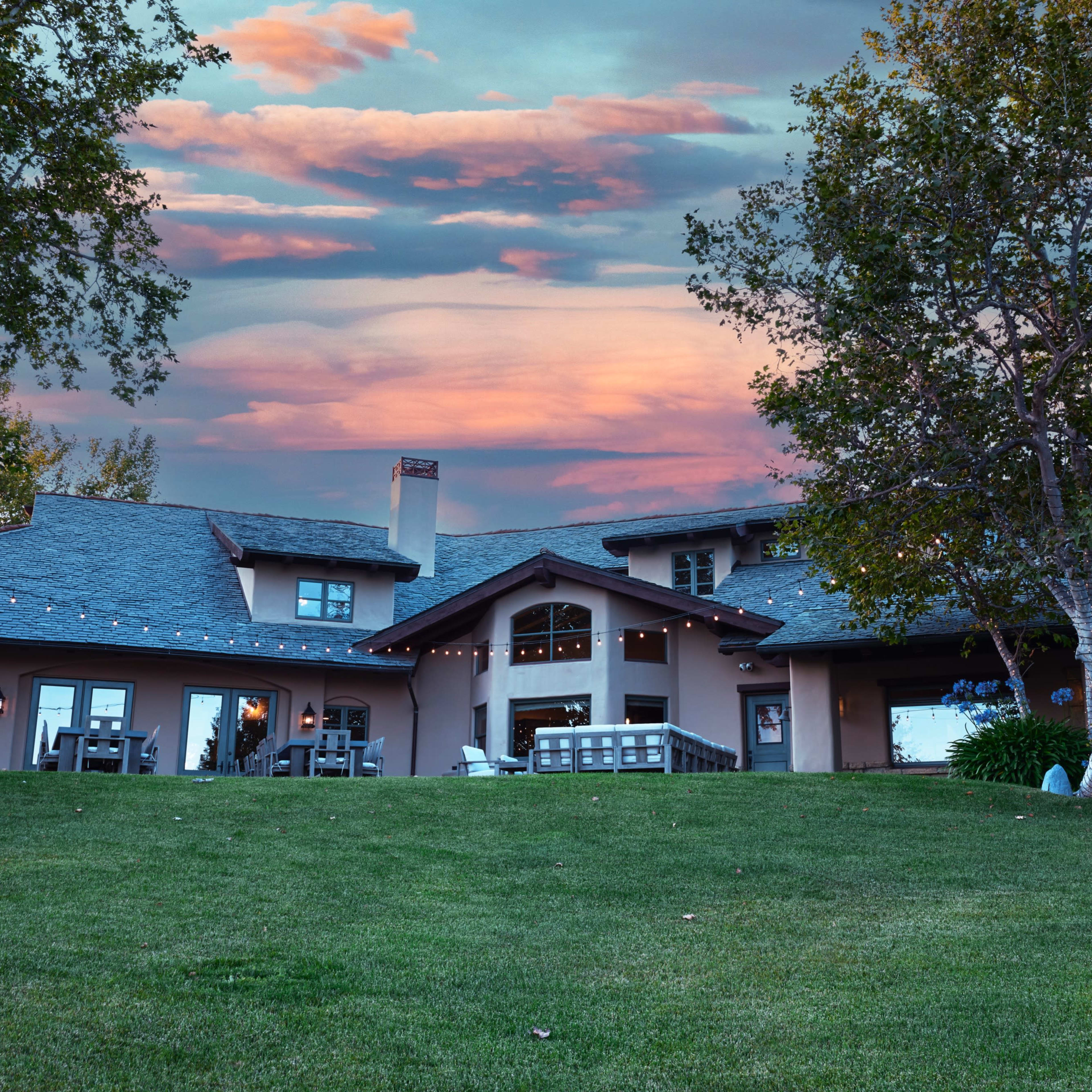 A spacious house with a large lawn, accompanied by a tree, against a backdrop of a colorful sky at sunset.