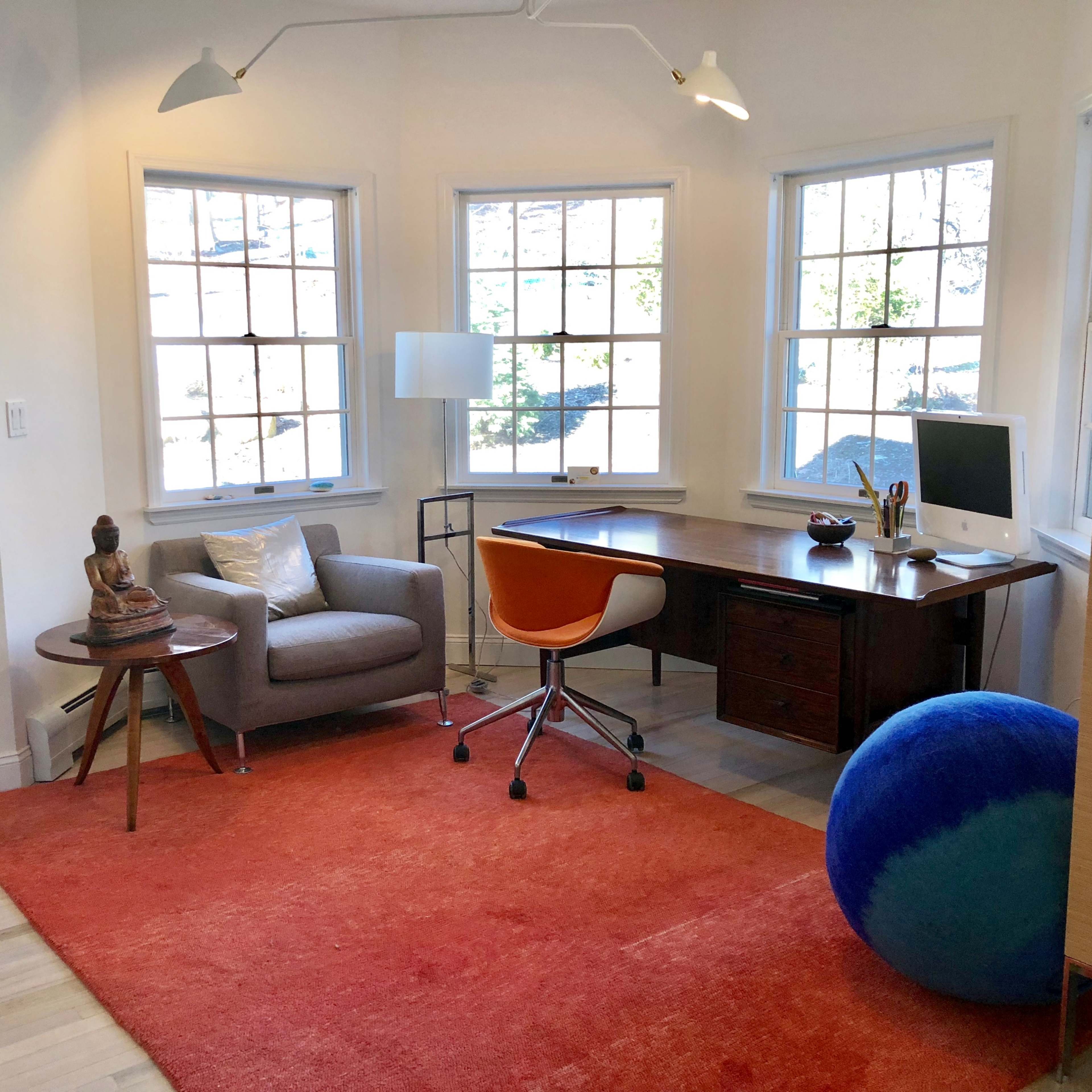 A bright home office features a wooden desk with an iMac, an orange chair, a gray armchair, a blue exercise ball, and large windows letting in natural light.