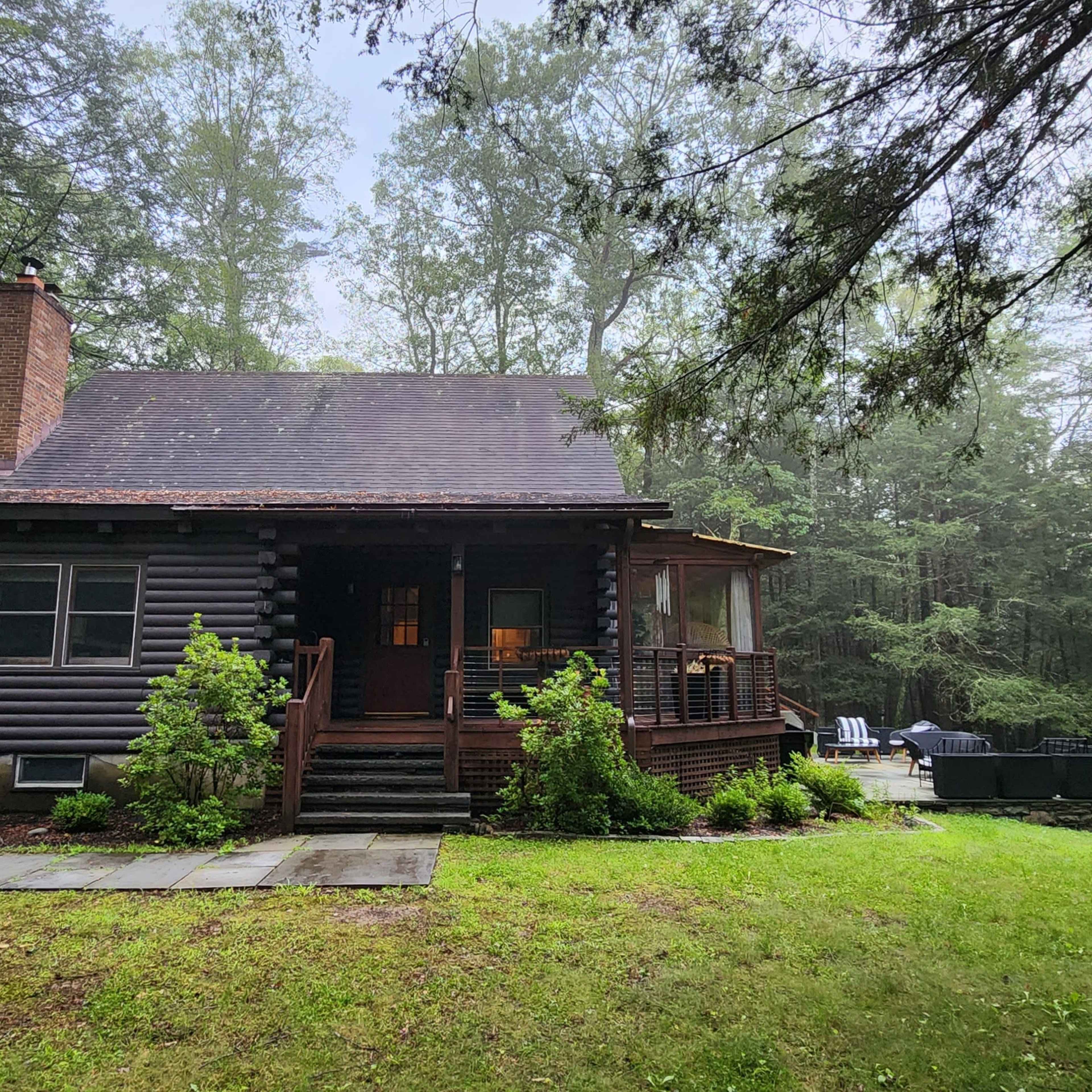 A log cabin is situated in a wooded area with green grass and stone walkways leading to its entrance.