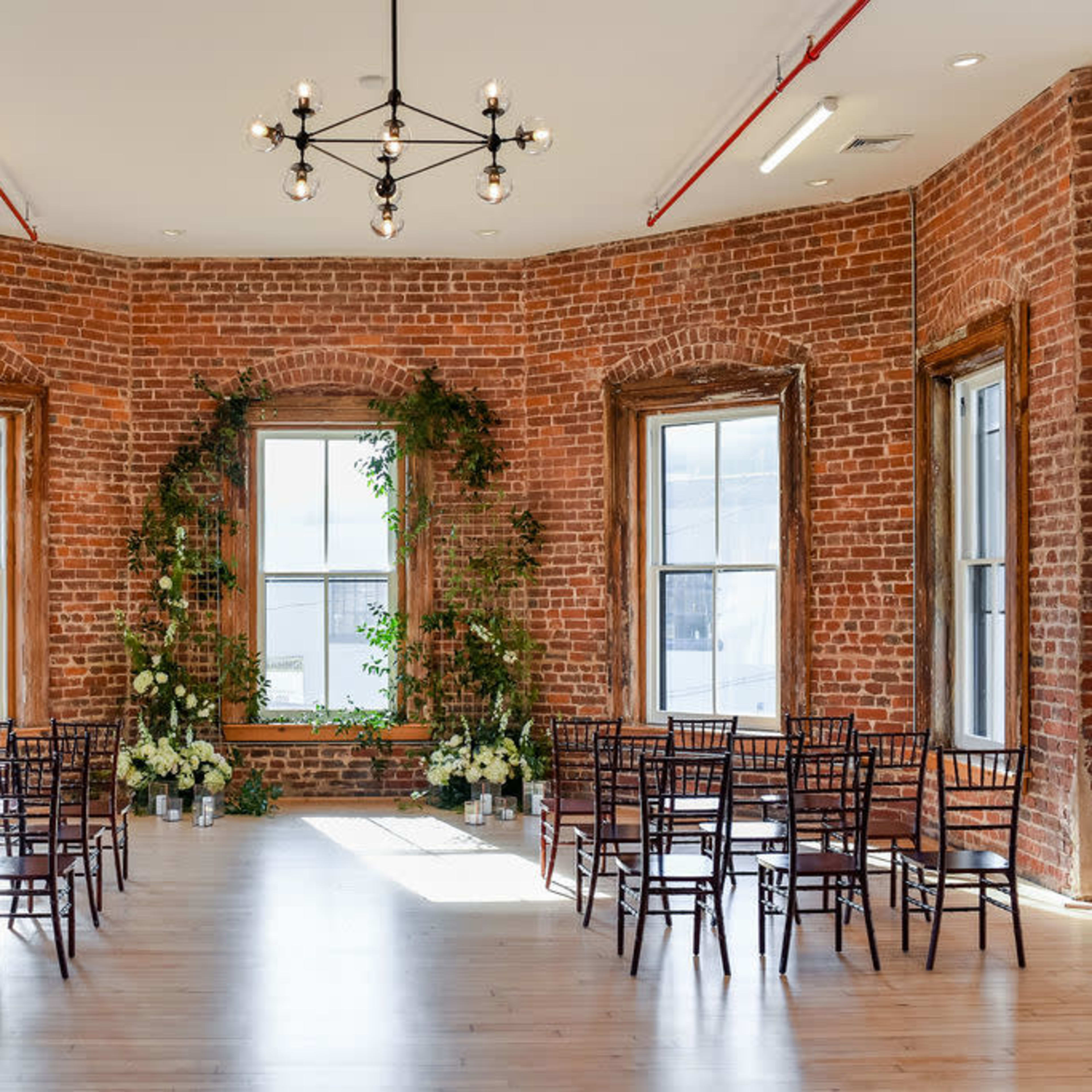 A room with exposed brick walls, adorned with plants and floral arrangements, featuring a circular seating arrangement of dark wooden chairs.