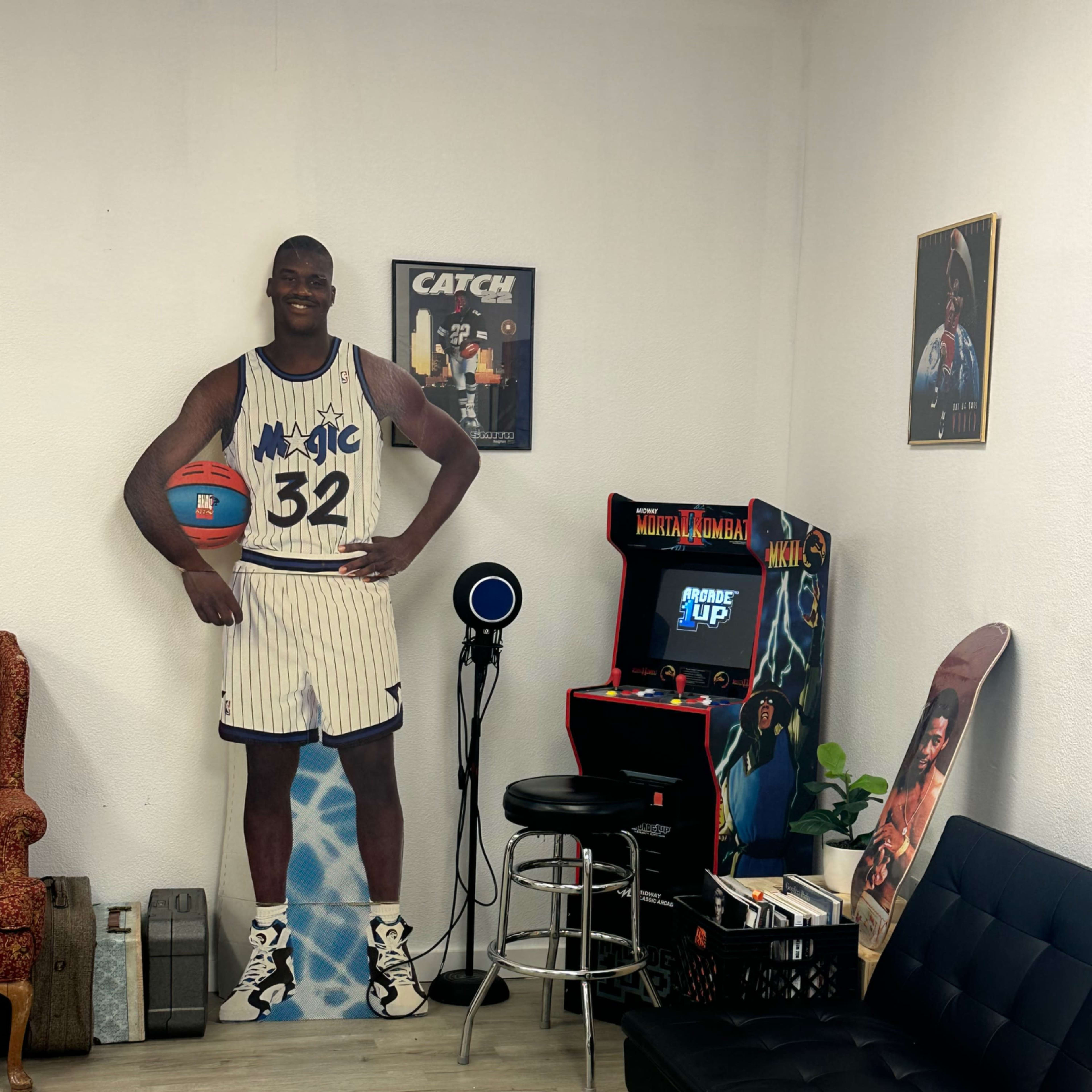 The scene features a life-sized cardboard cutout of a basketball player next to a vintage arcade machine in a recreational room with seating.
