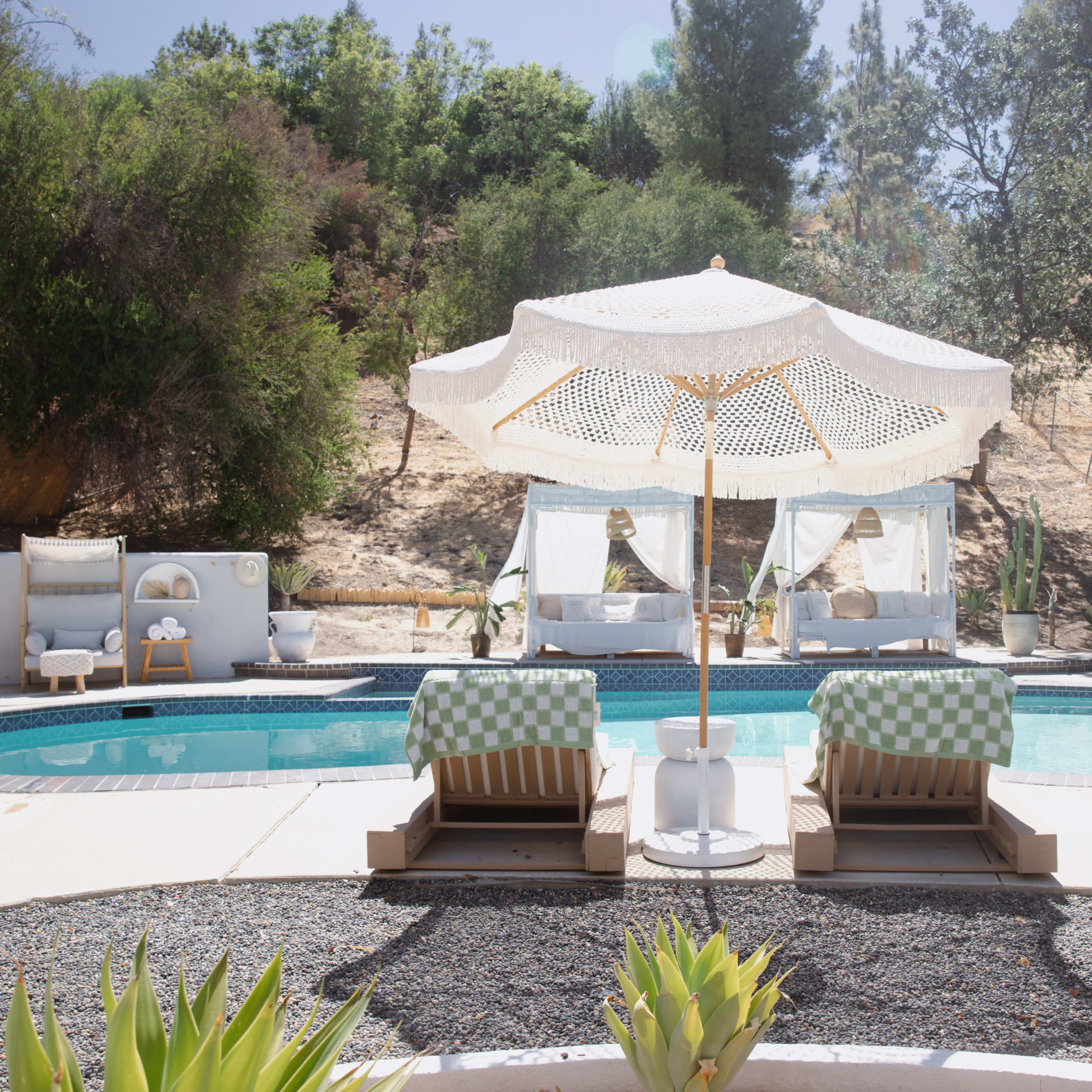 A swimming pool surrounded by lounge chairs and an umbrella, with cabana-style seating in the background and greenery on the hillside.