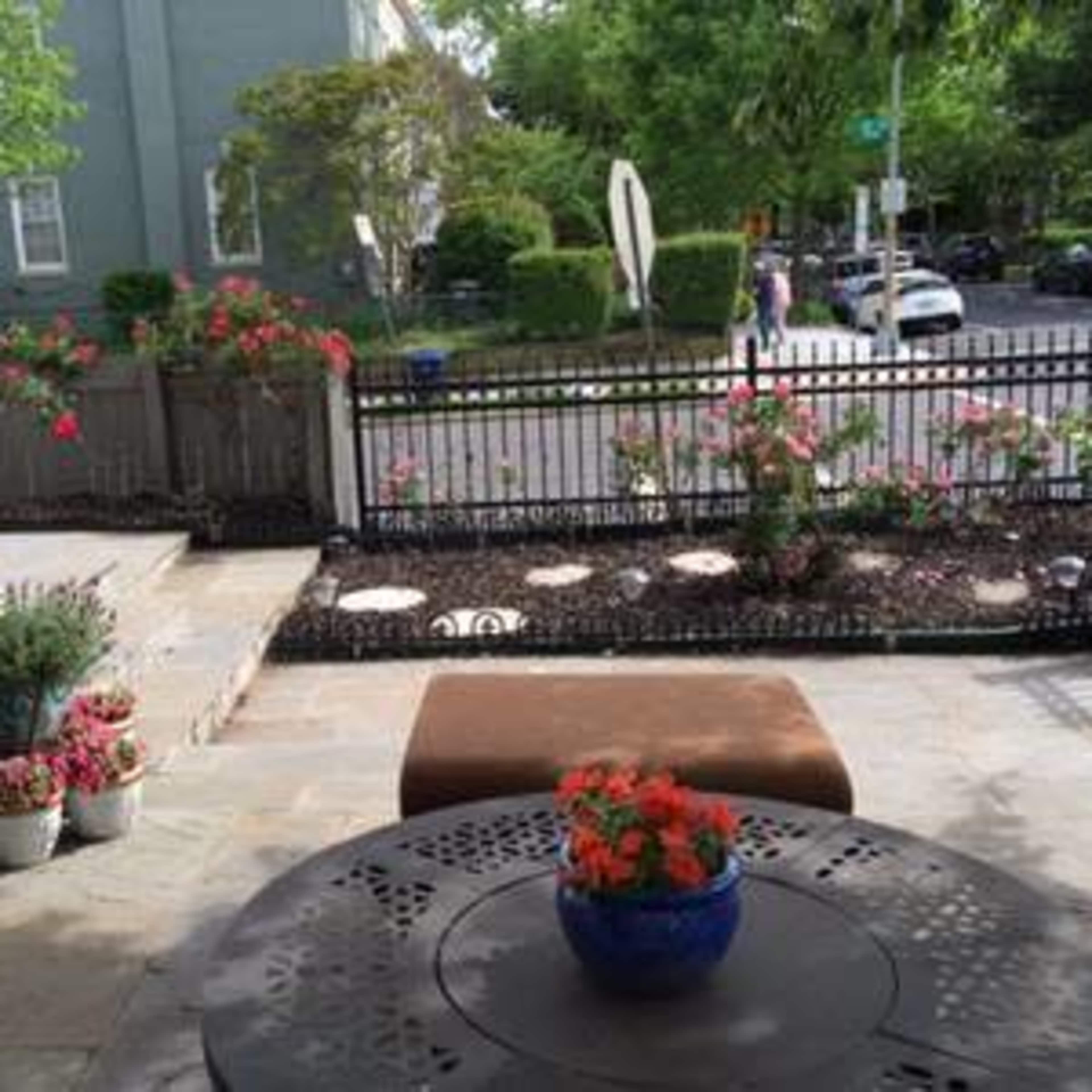 A patio area with a black metal table and decorative potted plants overlooks a garden space and street.