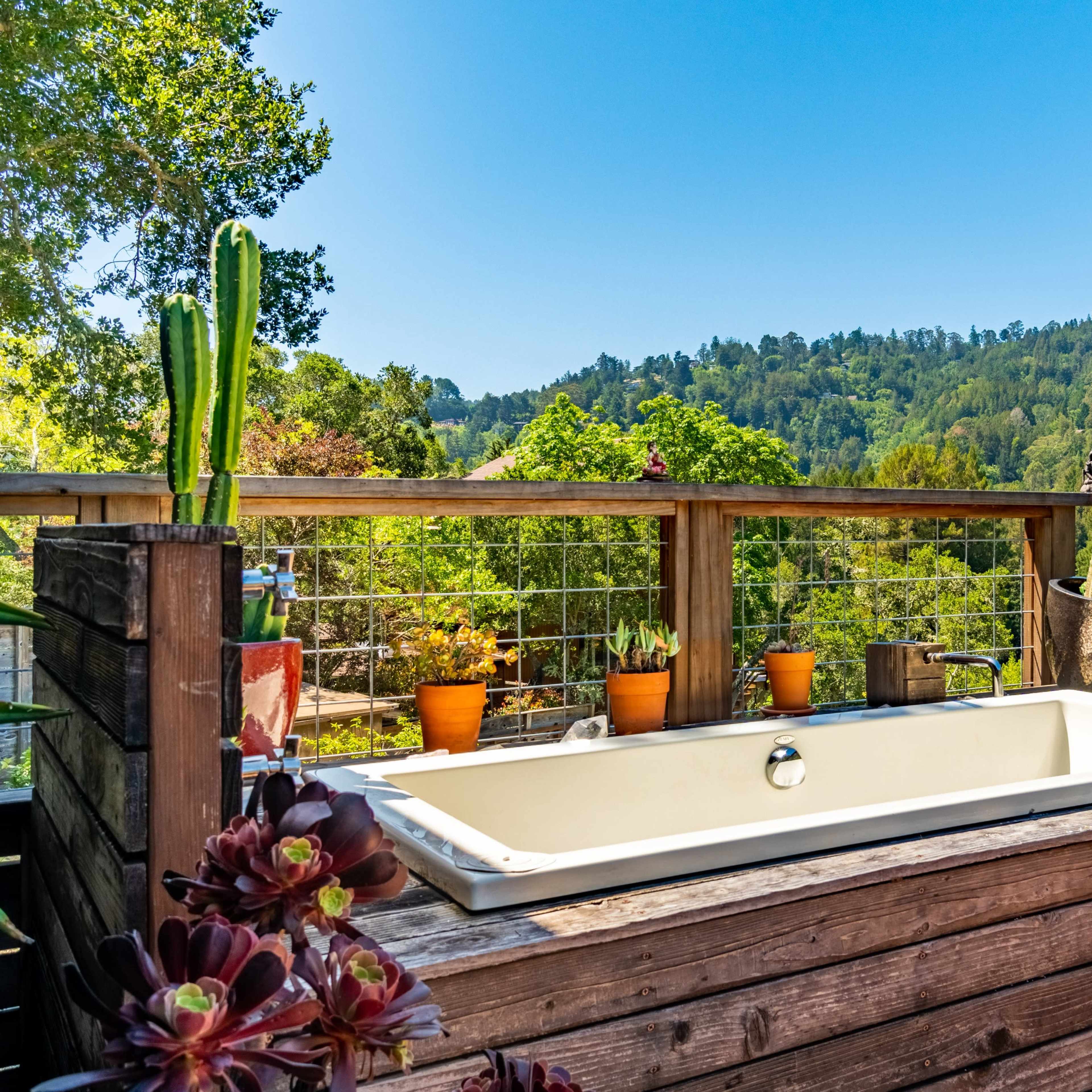 A wooden deck features a bathtub surrounded by potted plants and views of a green hillside.