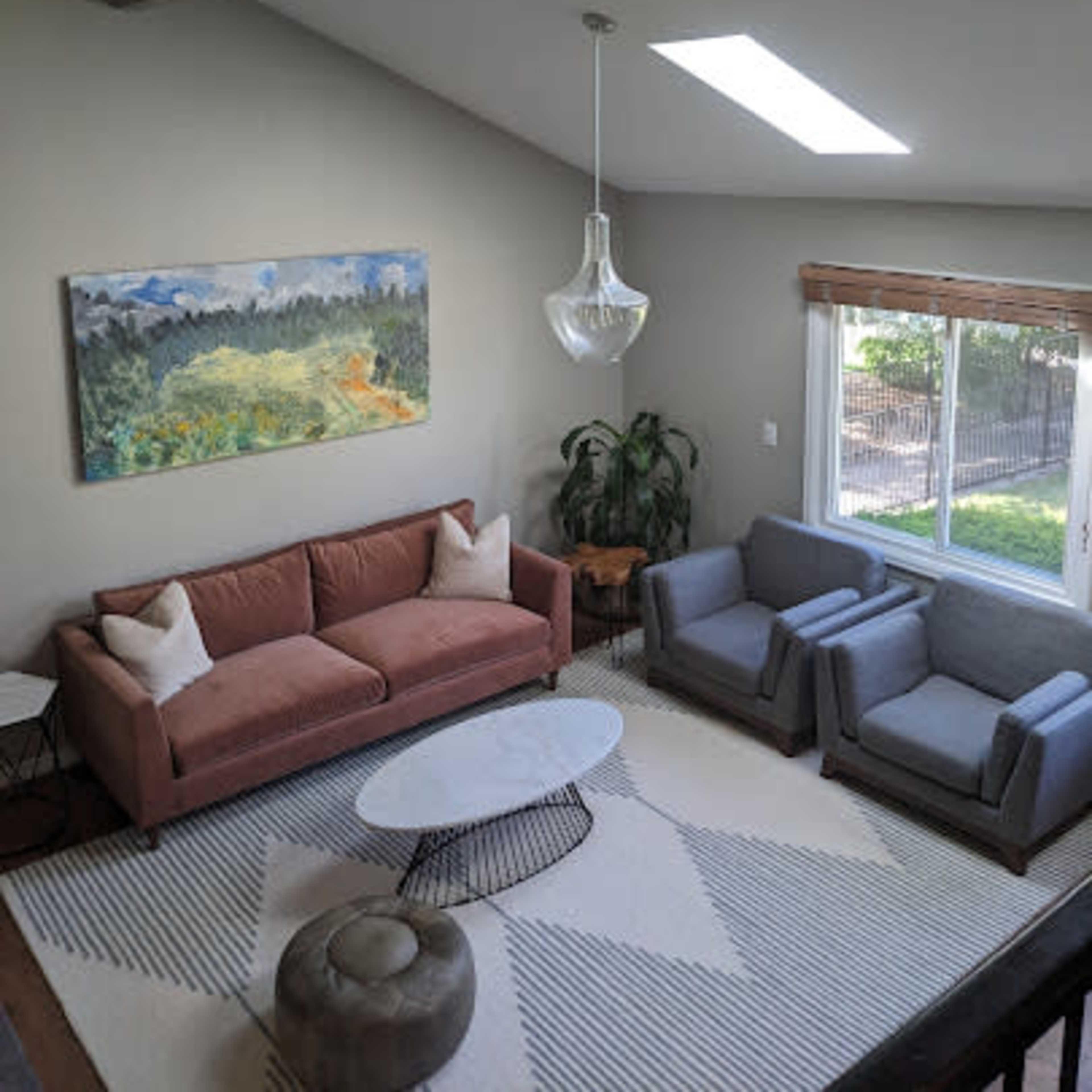 A modern living room with a pink sofa, two gray armchairs, a round coffee table, and a large window letting in natural light.