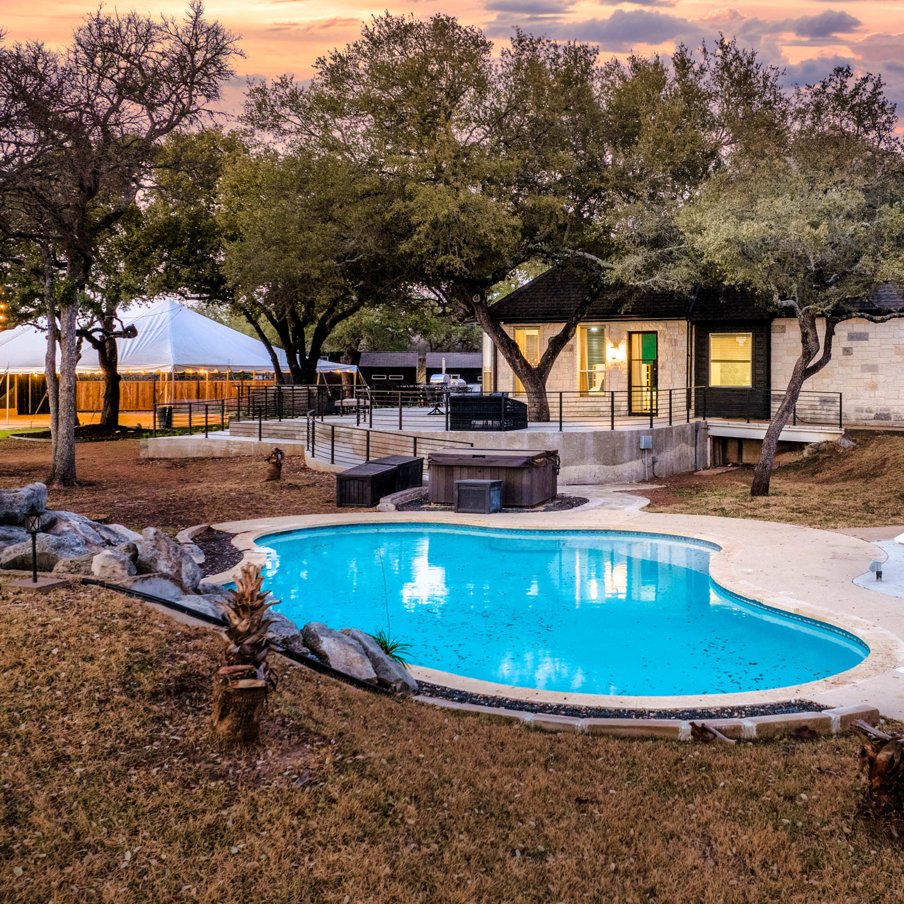 A residential property featuring a swimming pool with white lounge chairs, surrounded by grassy areas and trees, and a large tent in the background.