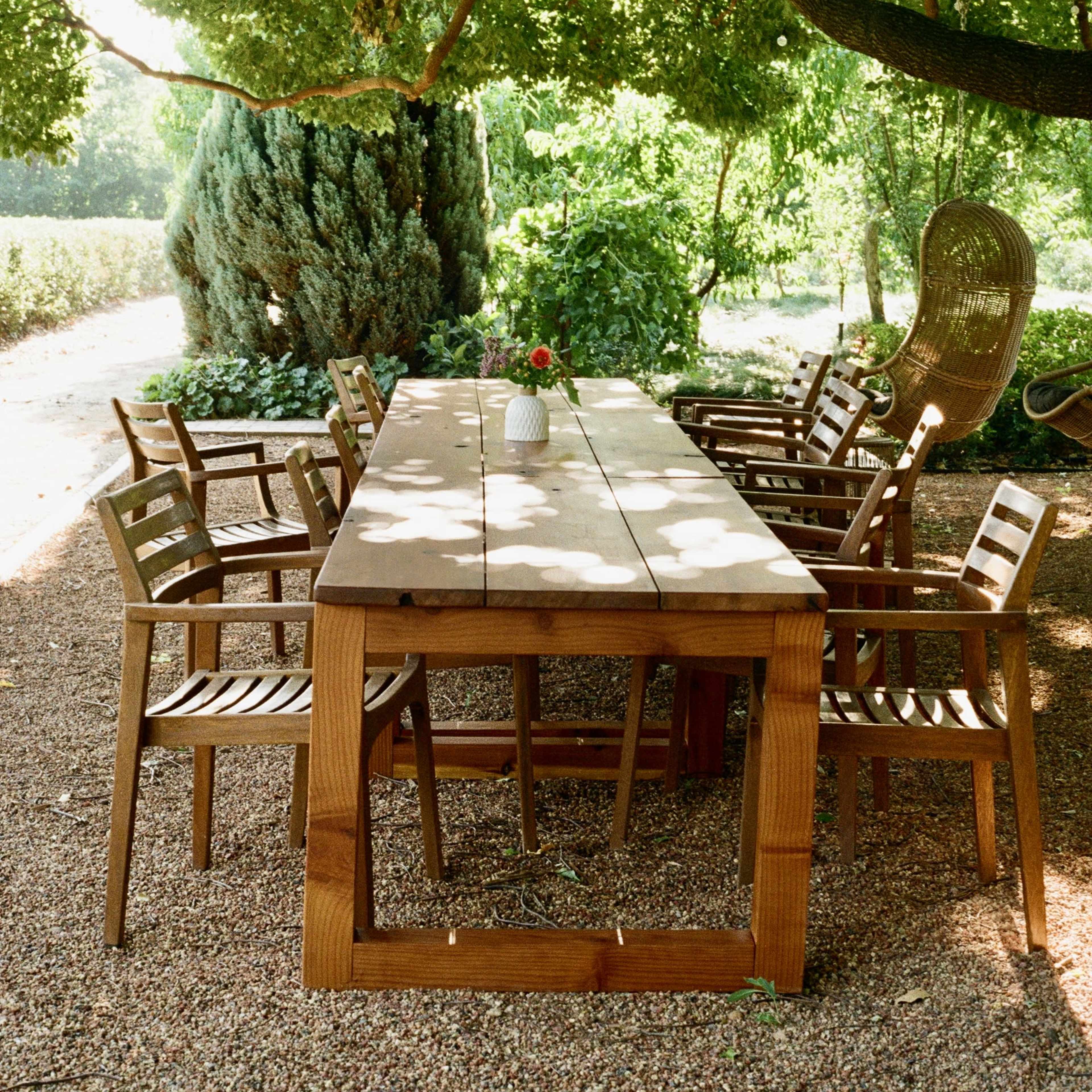 A wooden dining table surrounded by chairs is set under a lush tree in a garden.